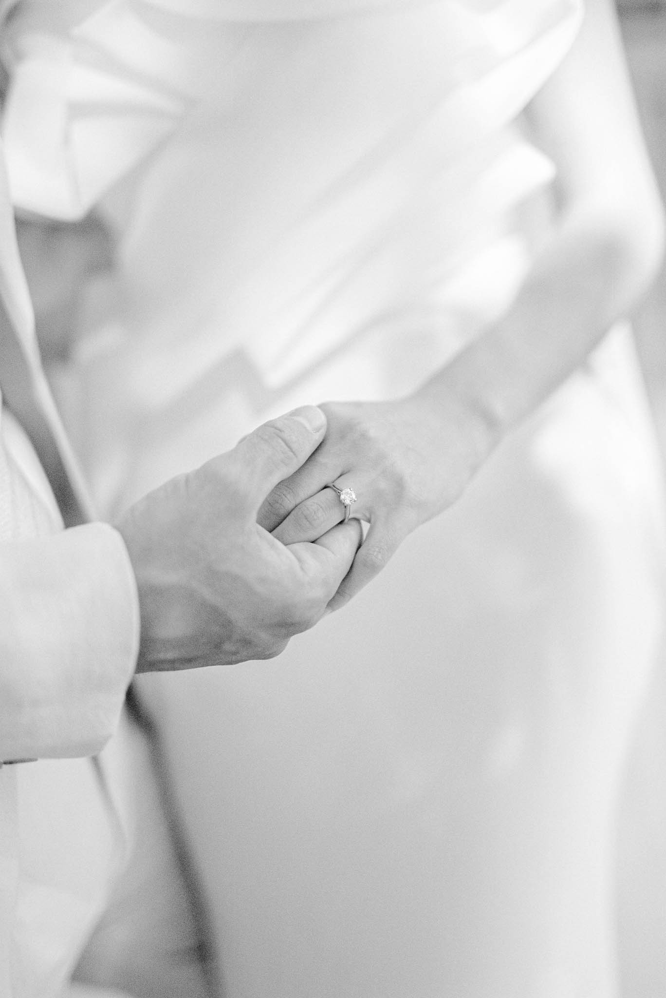 A black-and-white close-up detail shot of two people holding hands, with a solitaire diamond engagement ring visible on the bride's finger. The bride appears to be wearing a smooth, fitted white gown, and the second person — likely the groom — is wearing a light-colored suit jacket. The image is softly lit with bright highlights and gentle contrast, giving it an airy, high-key tone. The tight crop focuses entirely on the hands and ring, with the fabric of both outfits blurred in the background.