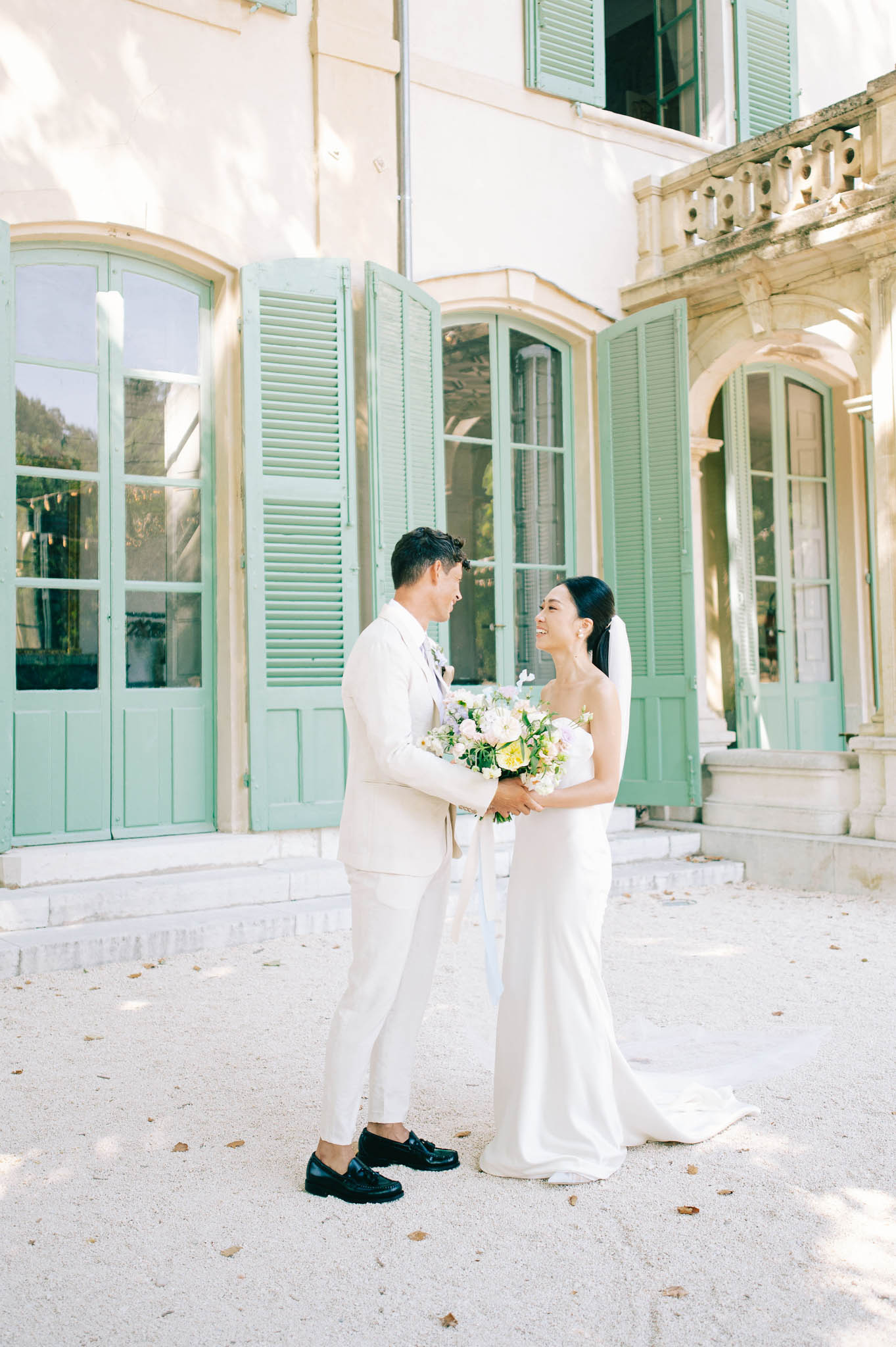 Bride in strapless white gown and groom in cream suit hold hands in courtyard of French bastide with green shutters