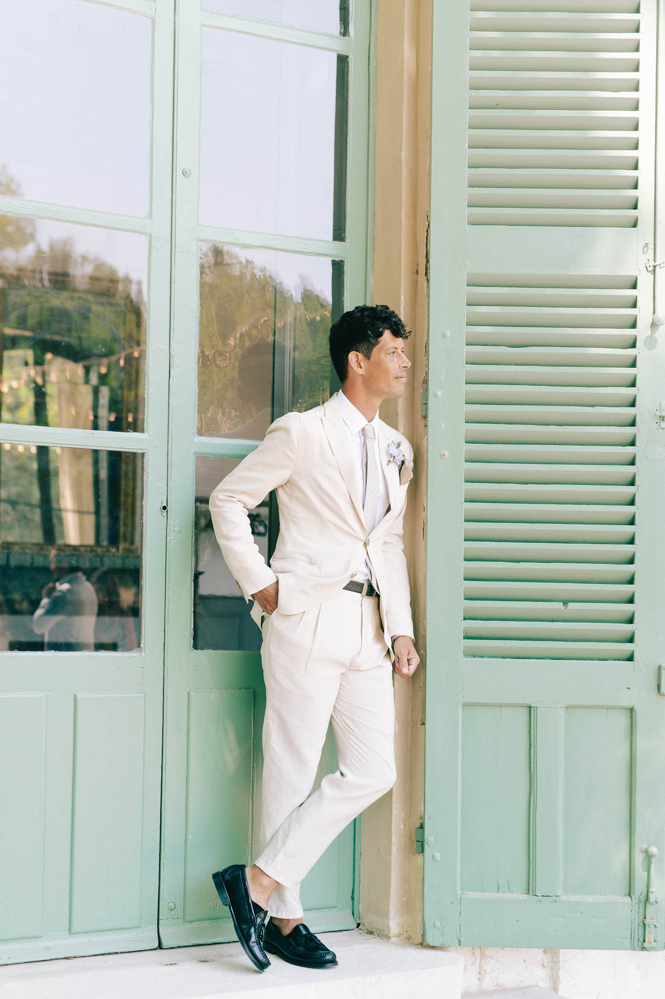 Groom in cream linen suit with lavender tie leaning against doorframe with sage green shutters at French venue