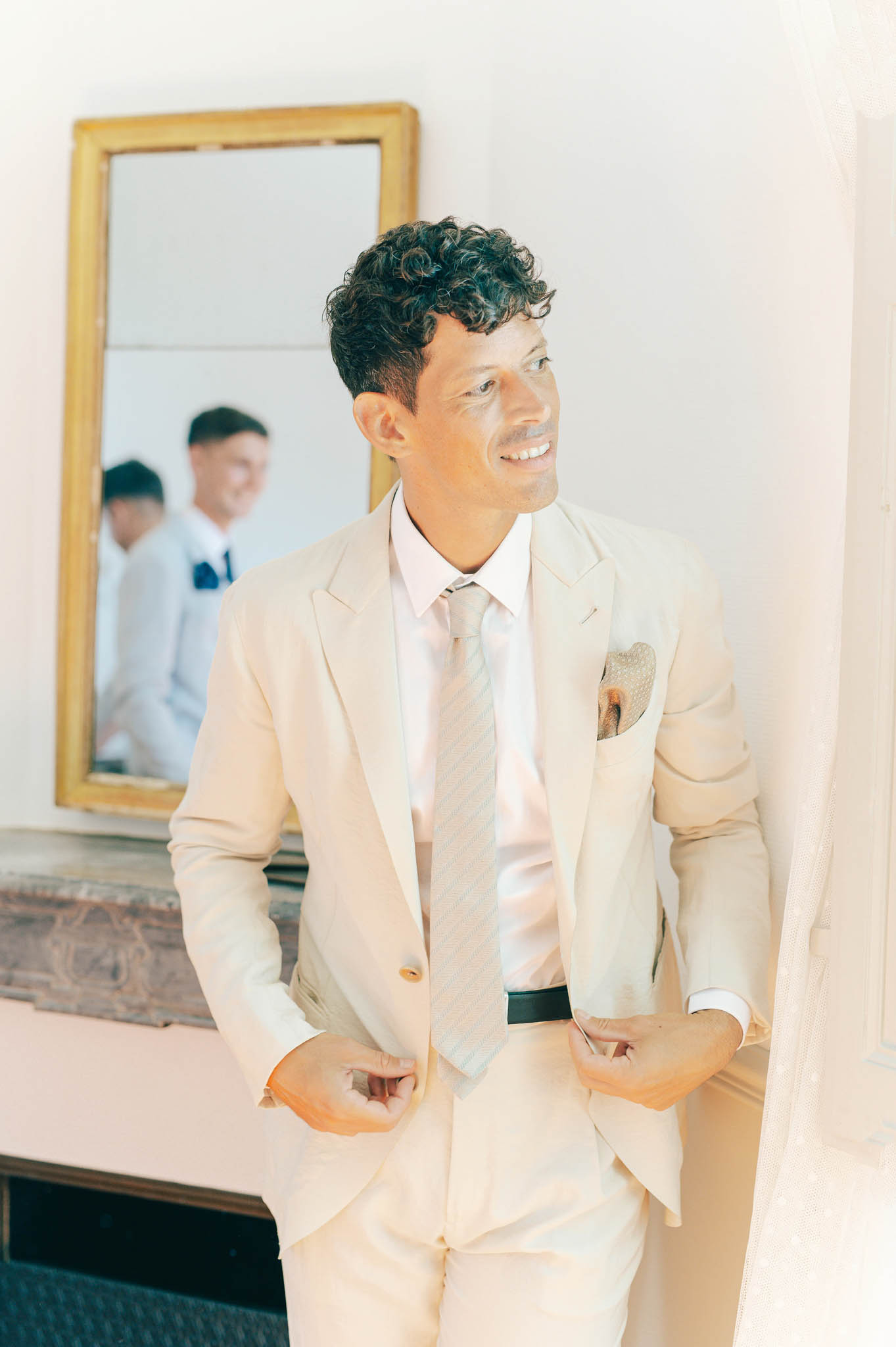 Groom in cream suit and pink shirt adjusting jacket with groomsmen reflected in gold mirror