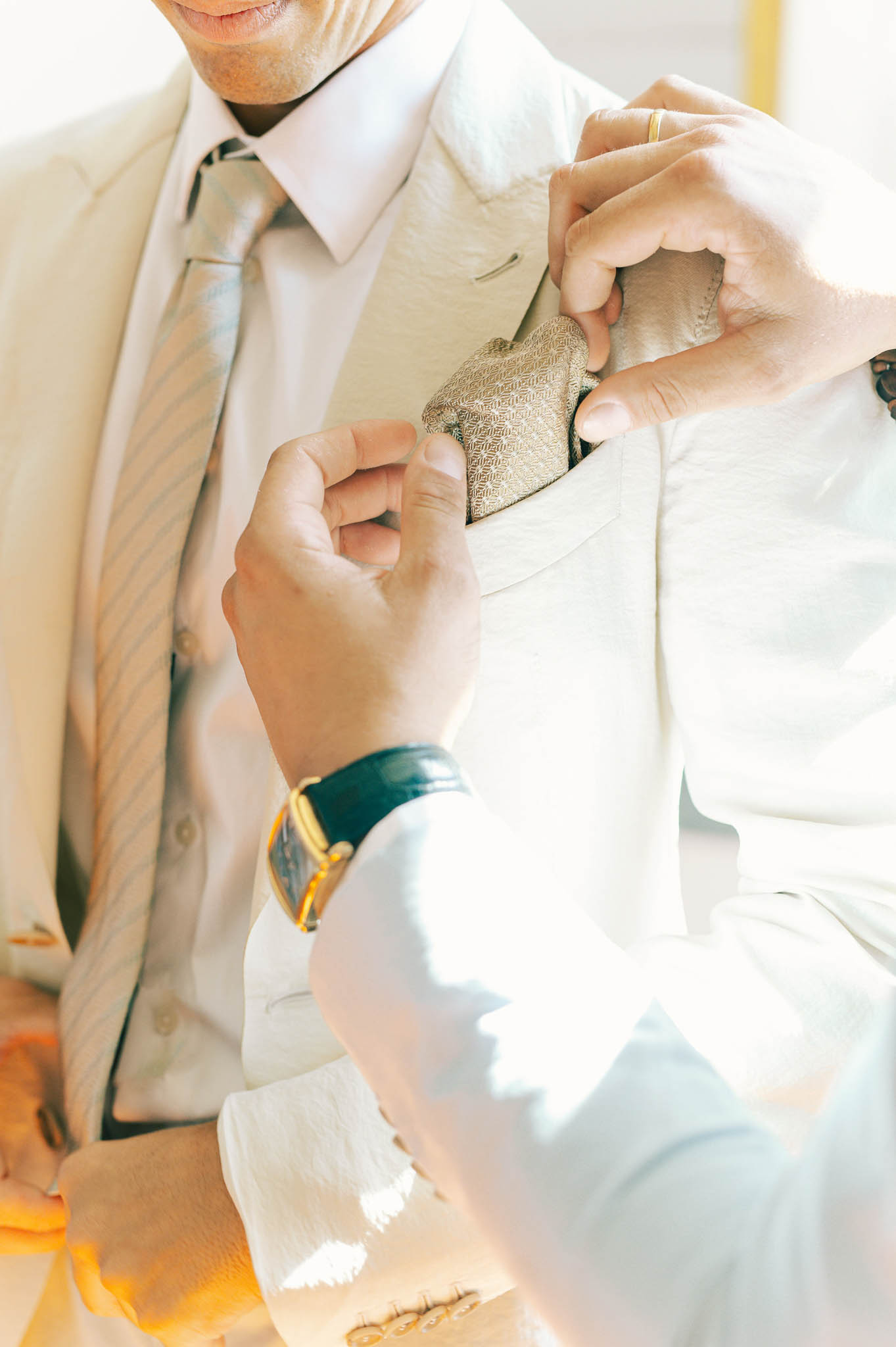Close-up of hands adjusting sage green pocket square into beige linen suit jacket