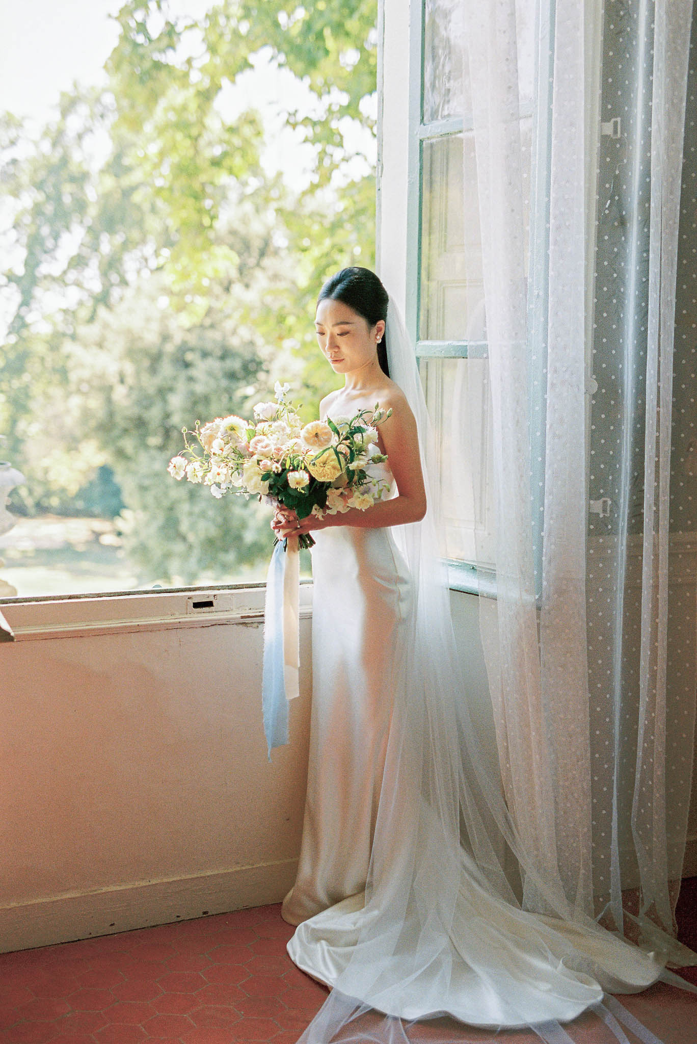 Bride in bias-cut satin gown with polka-dot veil holding garden bouquet by chateau window