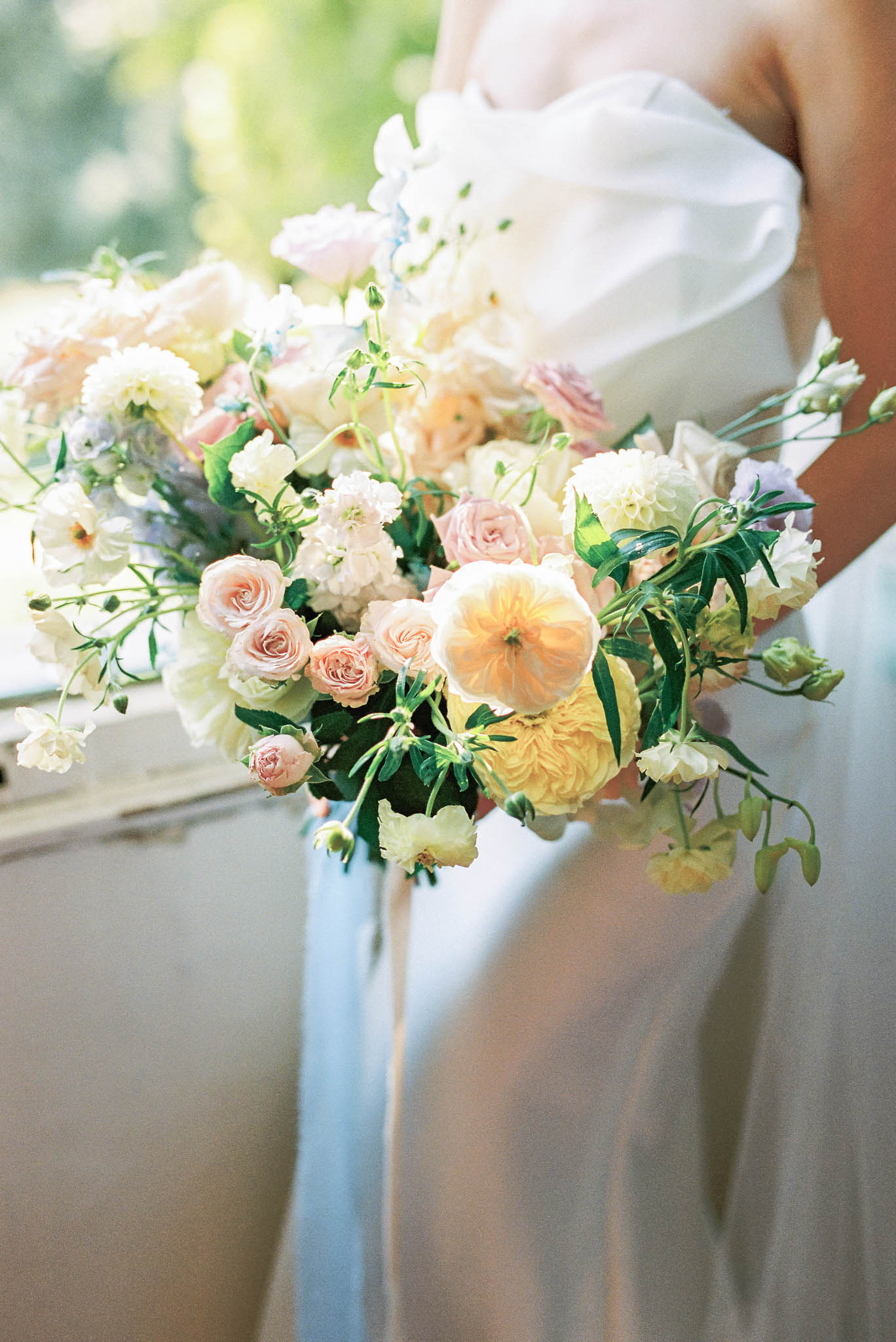 A close-up detail shot of a bride holding a large, loosely arranged bridal bouquet. The bouquet features garden roses in soft peach and blush tones, yellow David Austin-style roses, white carnations or lisianthus, pale lavender accents, and scattered greenery with trailing stems creating an unstructured, garden-gathered aesthetic. The bride is wearing a strapless ivory gown with a draped bodice, and long pale blue silk ribbons hang from the bouquet stems. The image is shot with a shallow depth of field, with the bouquet as the sharp focal point and the background softly blurred.