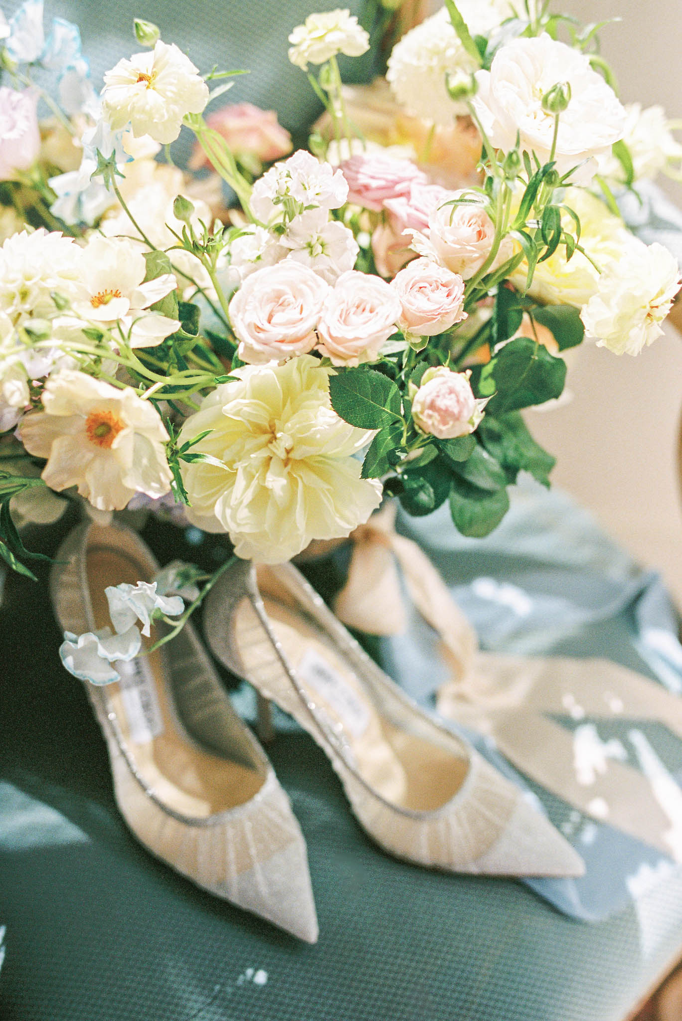 Bridal flat lay with ivory lace heels and pastel bouquet of blush roses, yellow dahlias, and blue sweet peas