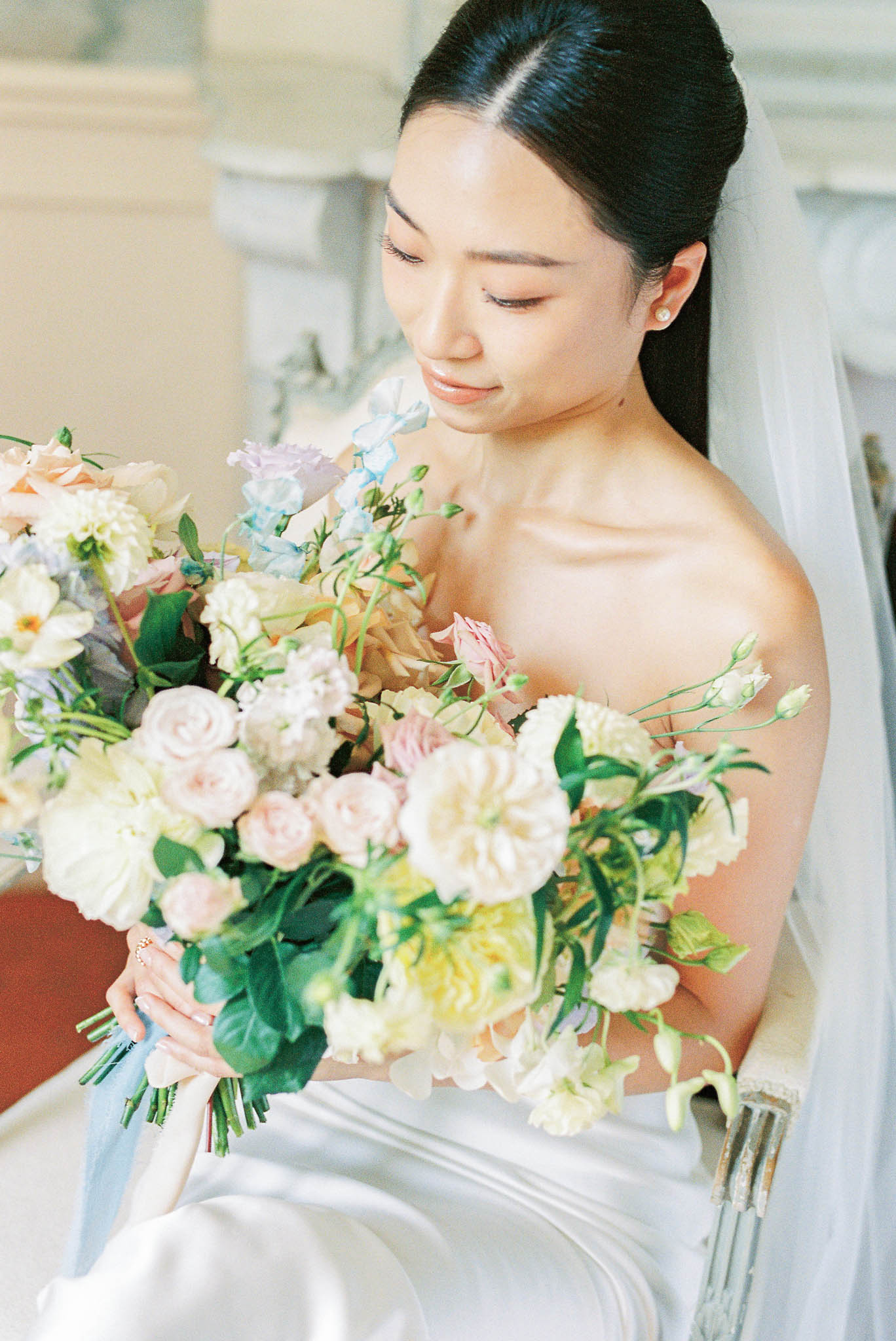 A close-up bridal portrait of a bride seated indoors, looking down at her bouquet. She wears a strapless ivory satin gown, a sheer veil with a soft edge, pearl stud earrings, and her dark hair pulled back in a sleek updo. The oversized bouquet is composed of blush garden roses, cream and yellow dahlias, pale pink lisianthus, blue and lavender sweet peas, cream carnations, and green foliage, creating a soft pastel palette with an organic, loosely structured arrangement. The background features ornate pale stone architectural detailing, suggesting a classic French château or hôtel particulier interior. The composition is a medium portrait shot with a shallow depth of field, with warm, diffused natural light.