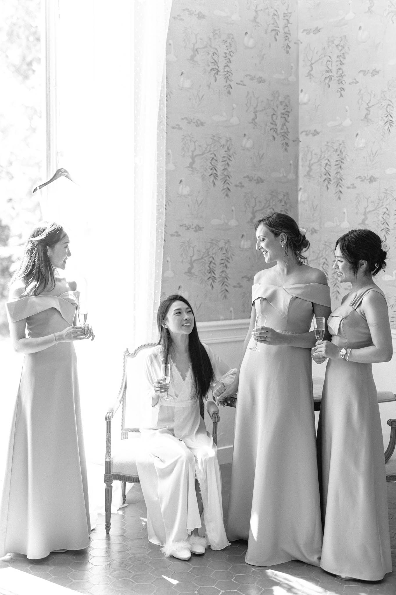 A black-and-white getting-ready scene showing a bride seated on a carved French-style chair wearing a silk robe and wide-leg trousers with fluffy slippers, holding a champagne flute and laughing with three bridesmaids. The three bridesmaids are dressed in floor-length off-the-shoulder satin gowns with a clean, minimal silhouette, and each holds a champagne flute. The group of four women are gathered near a large arched window with sheer curtains flooding the scene with bright natural light. The background wall features a decorative chinoiserie-style mural depicting swans, willow branches, and birds, rendered in soft tones against a light ground. The floor shows hexagonal tile work. The image is shot in bright, high-key black and white with soft contrast, captured as a candid mid-length portrait in an ornate interior room.