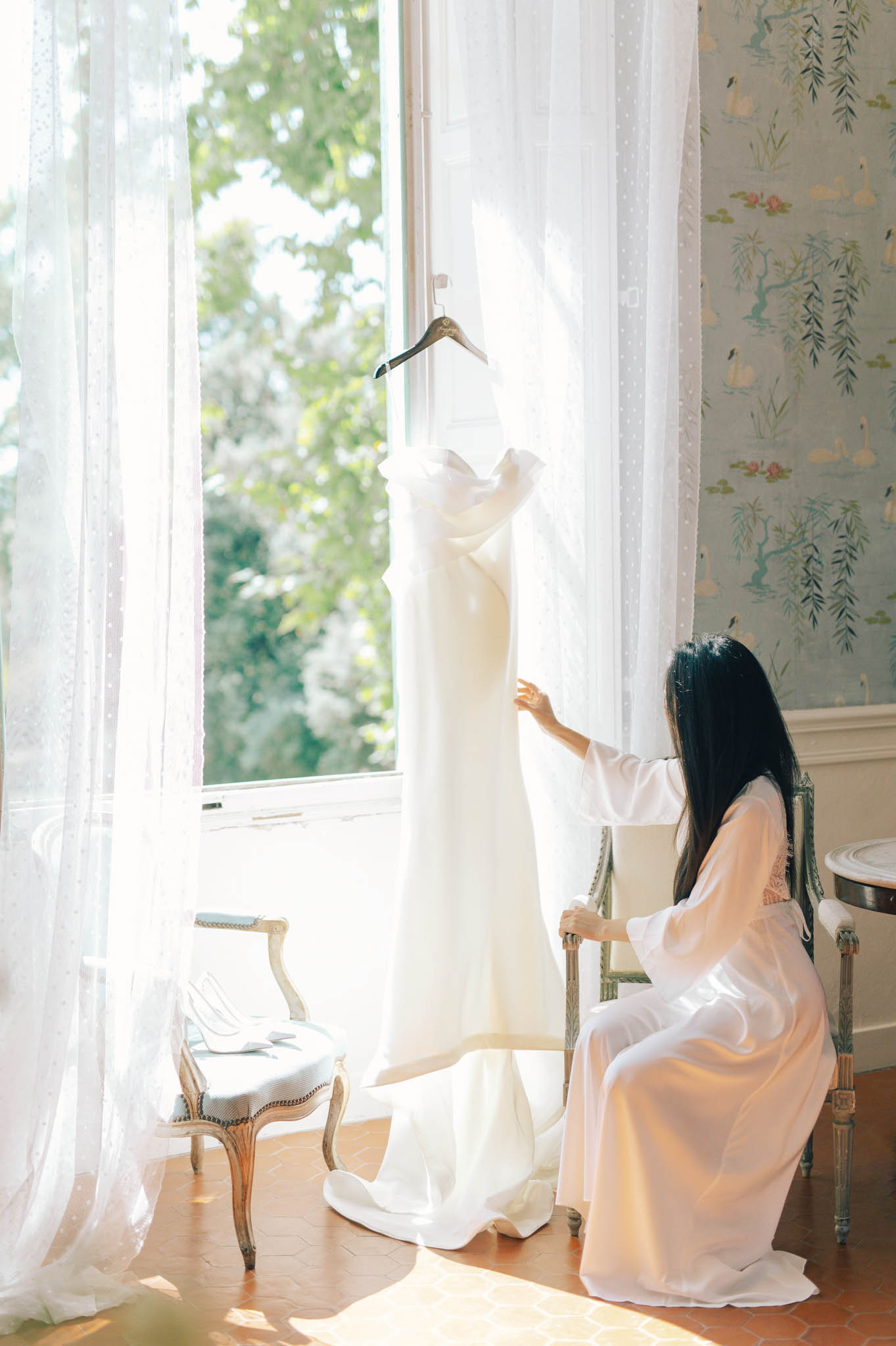 A bride in a white silk robe with long dark hair sits on a gilded French Louis XVI-style chair, reaching out to touch her wedding gown, which is hanging from a window frame in natural light. The gown is ivory, sleek, and column-cut with a clean neckline, displayed against sheer white Swiss dot curtains. The room features classic French interior details including pale blue-green chinoiserie wallpaper with botanical and bird motifs, terracotta tile flooring, and a second gilded stool visible to the left. The composition is a medium portrait shot taken from behind the bride, framing her against the bright window light in a getting-ready scene.