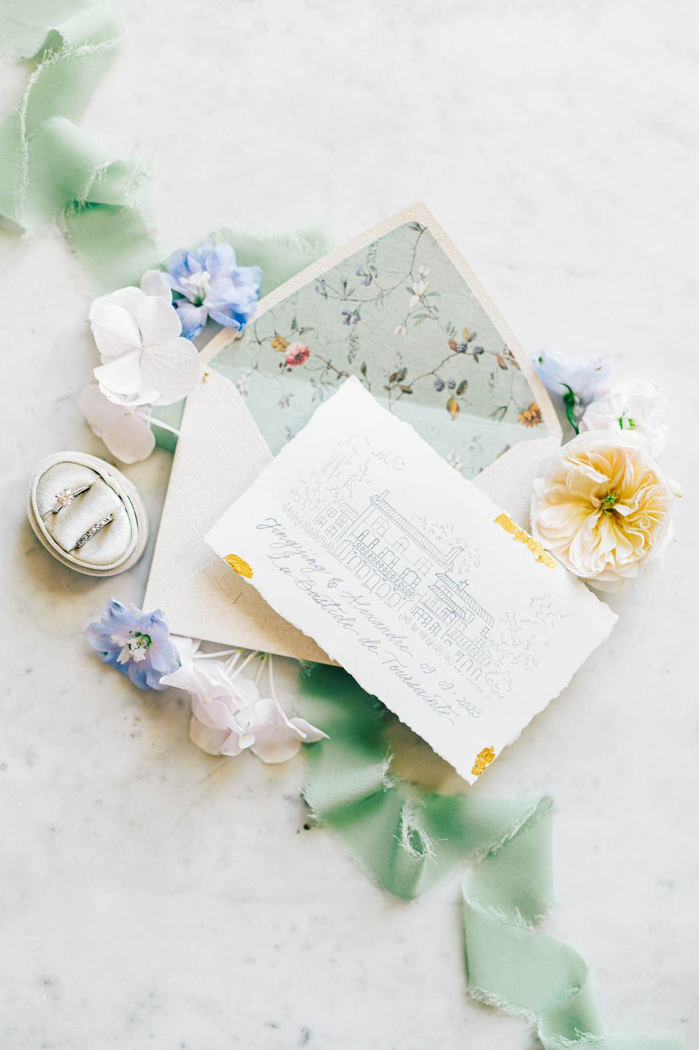 A flat lay detail shot of a wedding stationery suite arranged on a white marble surface. The invitation features deckled edges, hand-drawn architectural illustration of a venue, gold foil corner accents, and calligraphy script reading names and what appears to be 'La Bastide de Tourtour' with a date; it is paired with a cream linen-textured envelope lined with a sage green botanical floral print. Surrounding the stationery are loose blooms including a pale peach garden rose, blue and lavender delphinium florets, and white hydrangea petals, along with trailing mint green raw-edge silk ribbon. A velvet ring box in ivory holds two diamond rings positioned to the left of the suite.