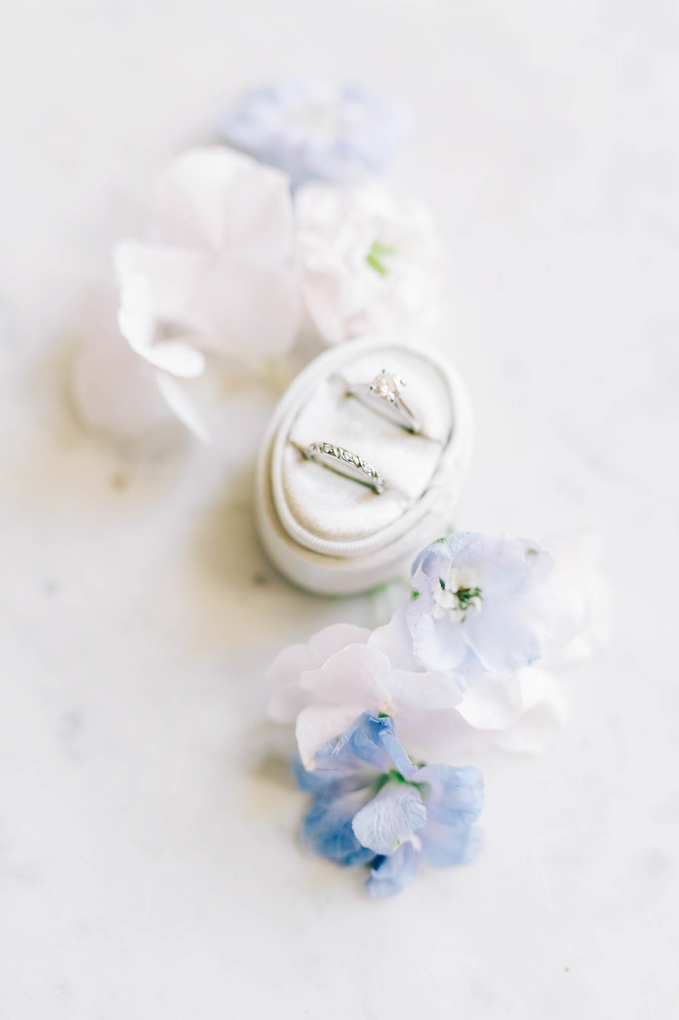 Diamond engagement ring and wedding band in ivory velvet ring box surrounded by blue sweet pea flowers on marble surface
