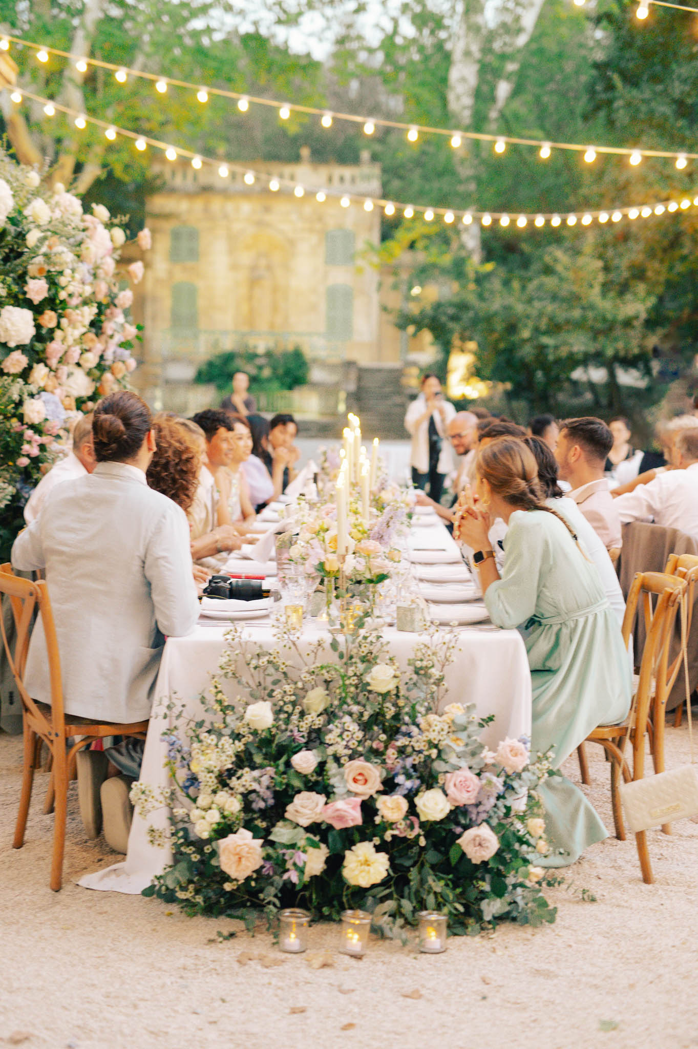 Long banquet table with blush and ivory rose runner, taper candles, and bistro lights in chateau courtyard