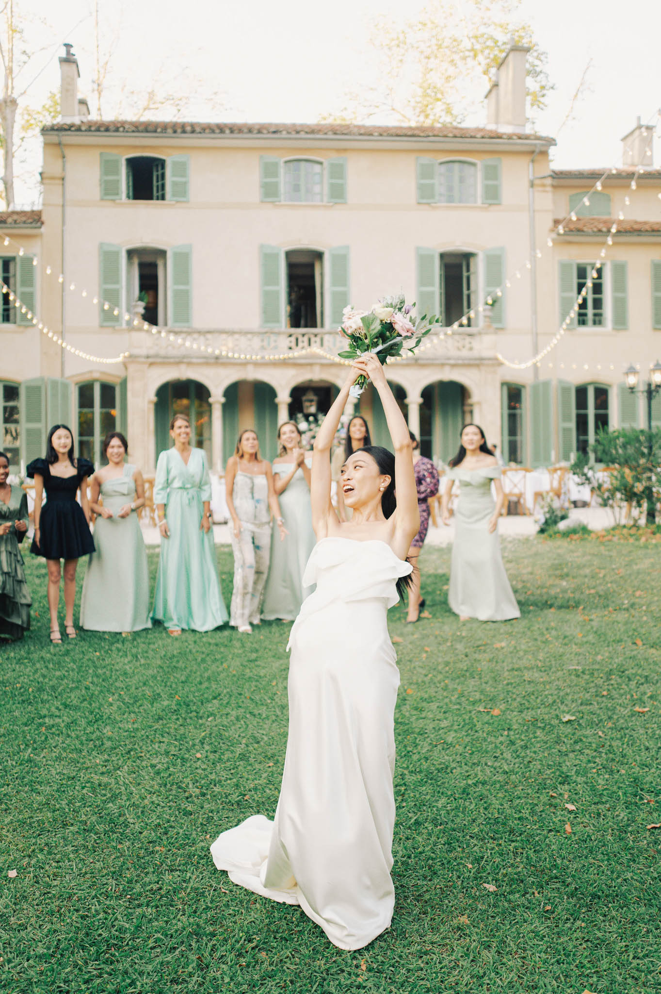 The bride is mid-bouquet toss on the lawn of a Provençal-style manor house, her arms raised overhead holding a small bouquet of blush pink and lavender flowers with greenery. She wears a white strapless gown with a structured ruffle detail at the neckline and a floor-length skirt with a slight train. Approximately eight female guests stand in a loose line behind her, dressed in a mix of outfits including a mint green jumpsuit, sage and seafoam gowns, a black mini dress, and casual separates, creating an eclectic, unstyled look among the group. The venue facade features warm ochre-toned render, sage green shutters, arched ground-floor openings, and a balcony, with bistro string lights draped across the foreground and reception furniture visible to the right. The shot is a full-length portrait taken from a low-to-mid angle, with the bride in sharp focus and the guests and building softly blurred in the background. Potential venue feature image.