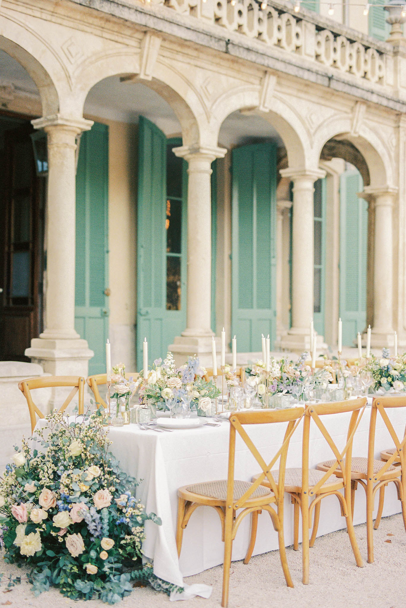 An outdoor wedding reception table is set in front of a classic French château facade featuring arched colonnades and distinctive sage-green shuttered doors. The long rectangular table is dressed in a white linen cloth and lined with natural wood cross-back chairs with cane seats. The centerpiece design runs the full length of the table, featuring clusters of blush roses, ivory blooms, soft blue delphinium, and eucalyptus foliage, with a large cascading floral arrangement spilling off the near end of the table onto the ground. Tall white taper candles in individual holders are placed at intervals along the center of the table, creating a repeated vertical element. Place settings with white plates and grey napkins are visible, along with glassware. The overall decor palette is soft and muted — blush, ivory, dusty blue, and green — with a classic French countryside styling. Wide shot composition. Potential venue feature image.