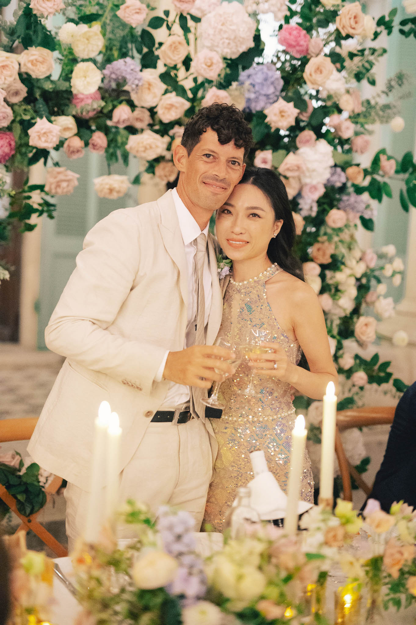 Bride in sequined mini dress and groom in cream suit before blush rose and lavender floral wall at reception