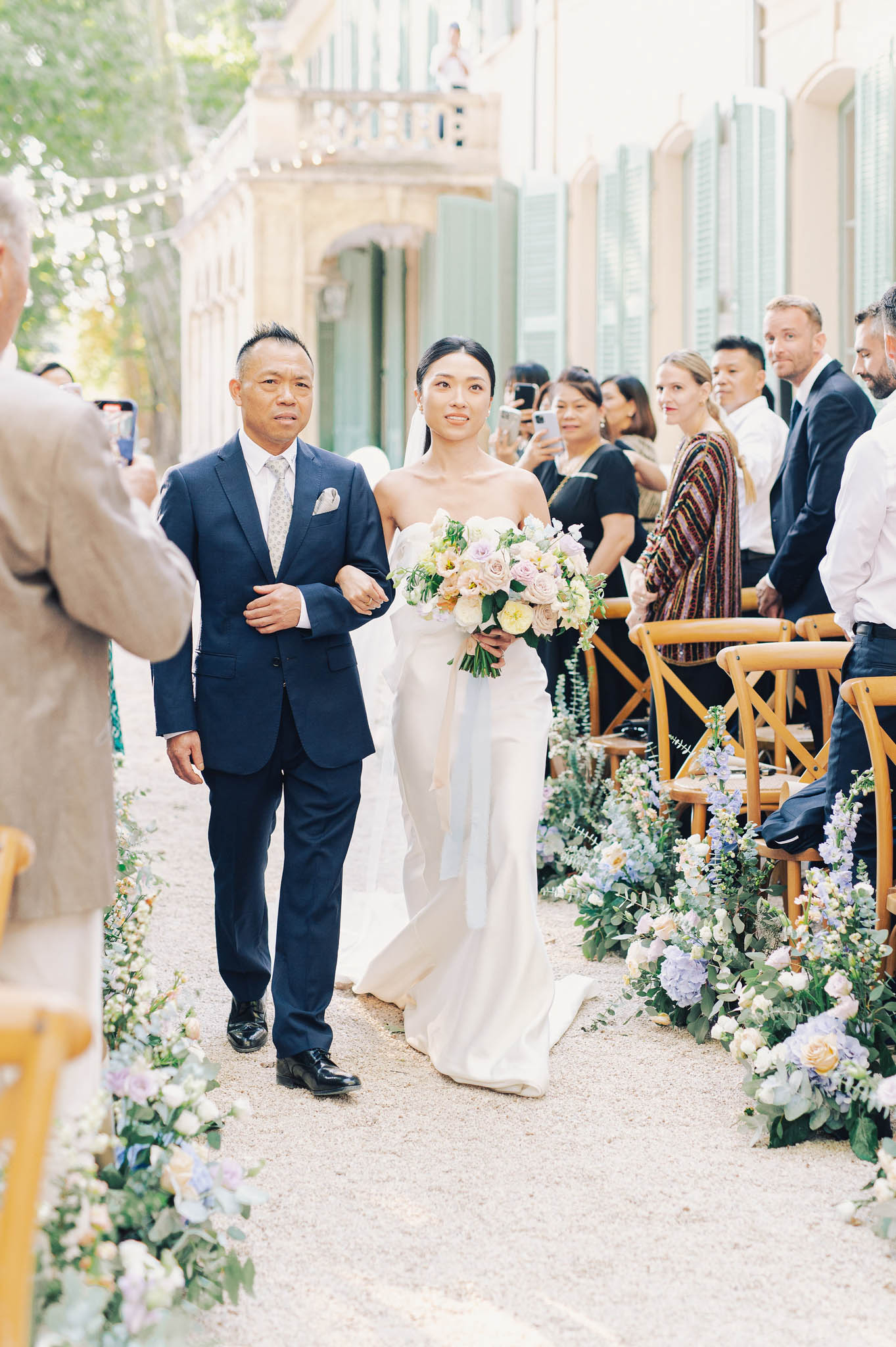 The bride is being walked down the aisle by a male escort, likely her father, during an outdoor ceremony at a French château or manor house with sage green shutters and a balcony visible in the background. The bride wears a strapless ivory fitted gown with a train and veil, and carries a loose bouquet featuring blush roses, lavender blooms, soft yellow flowers, and greenery. The aisle is lined on both sides with low floral arrangements of blue hydrangeas, ivory roses, peach garden roses, and trailing greenery, with natural wood cross-back chairs seating approximately 20 or more guests who are watching and photographing the procession. The shot is a medium wide portrait taken at ground level along the gravel aisle, capturing the couple walking toward the officiant with the historic façade of the venue as a backdrop.