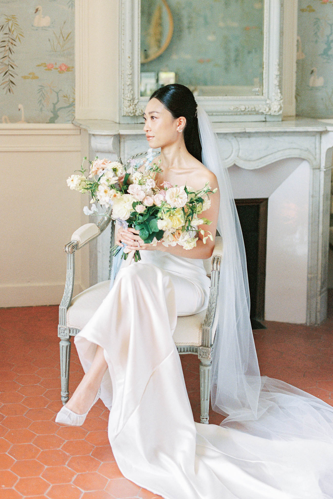 Bride seated in chateau room wearing strapless ivory satin gown and blue-grey veil holding blush and cream bouquet