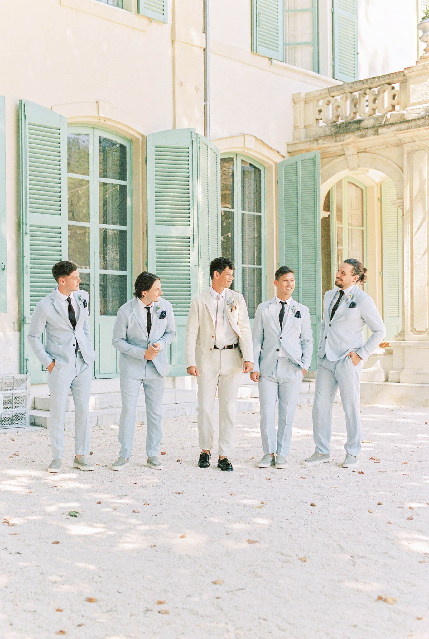 Groom in cream linen suit and four groomsmen in light blue suits walking across chateau gravel courtyard