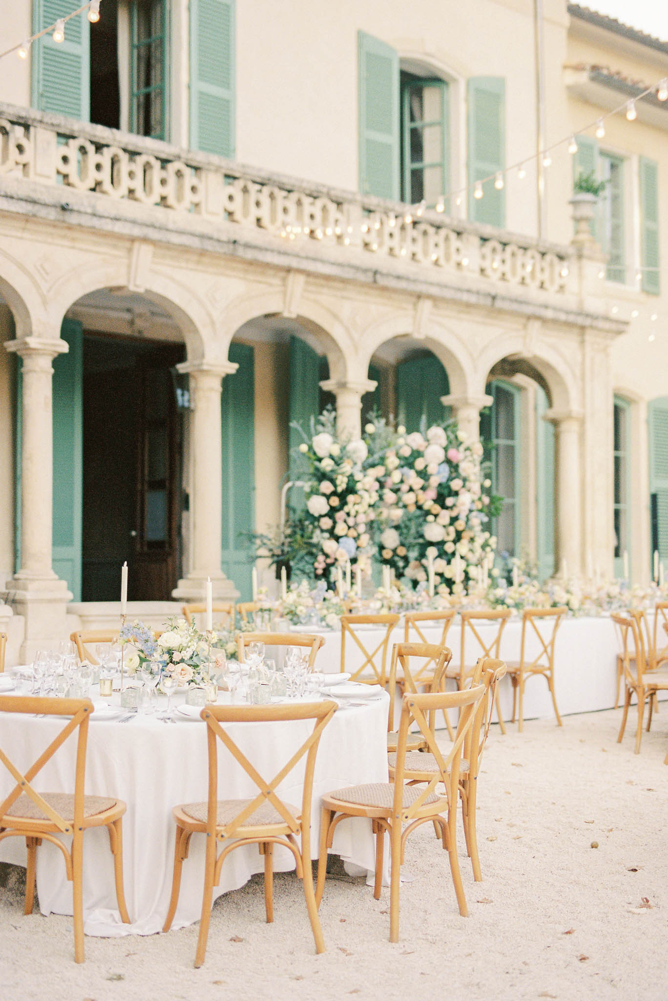 An outdoor wedding reception setup in the courtyard of a French manor or château, featuring cream-rendered stone architecture with teal-green shutters, arched colonnades, and an ornate stone balustrade on the upper level. Round guest tables are dressed in white linen and surrounded by natural wood cross-back chairs, set on a gravel surface; the foreground table is styled with low centerpieces of soft blue, blush, and ivory blooms with greenery, multiple wine glasses, and taper candles. Behind the guest tables, a long head table is visible, anchored by a large freestanding floral installation featuring ivory, blush, peach, and pale blue blooms mixed with lush greenery, flanked by tall white taper candles. Globe string lights are strung across the façade, adding warm ambient lighting to the early-evening scene. The overall decor palette is soft and pastel — white, blush, blue, and natural wood — with a classic French-country styling approach. Wide establishing shot. Potential venue feature image.