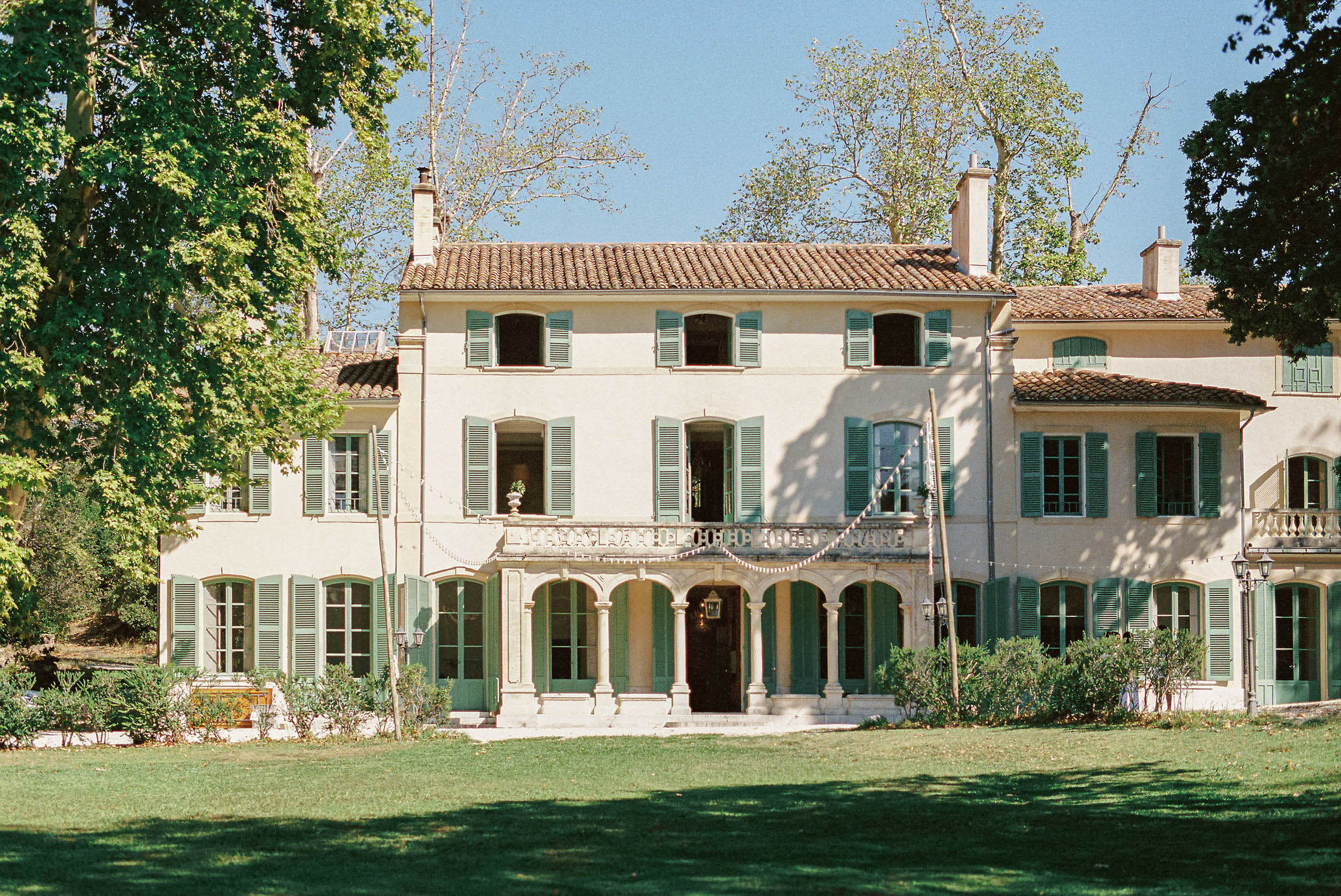 Provencal manor with cream walls, sage green shutters, arched colonnade, and fairy light bunting