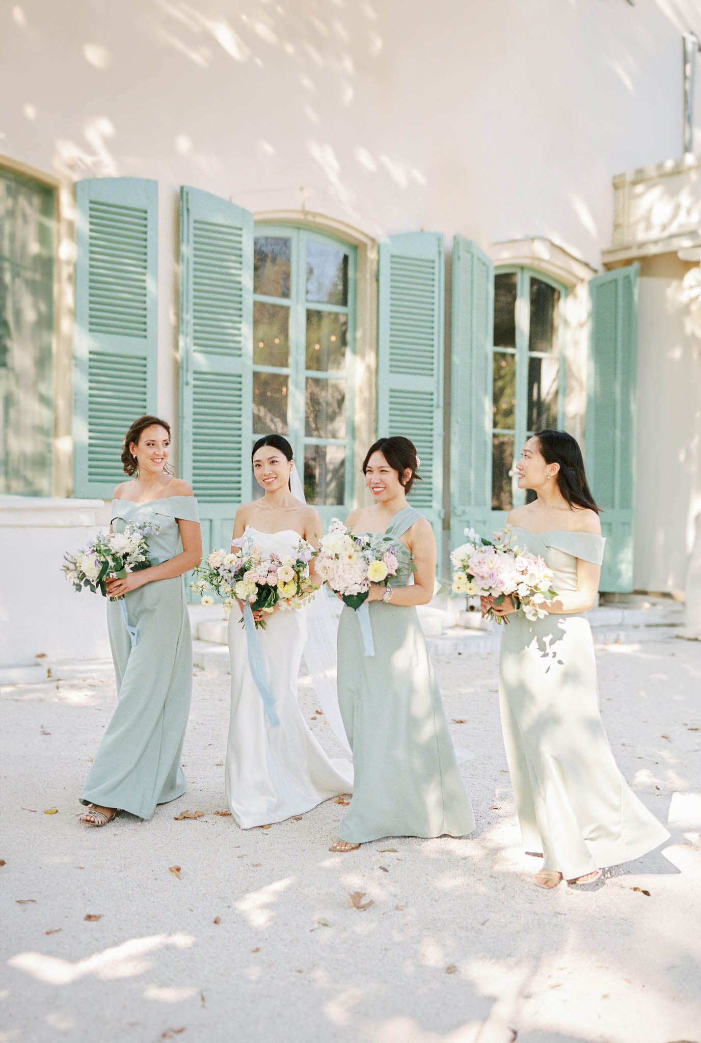 Bride with three bridesmaids in sage green gowns holding blush and lavender bouquets in chateau courtyard