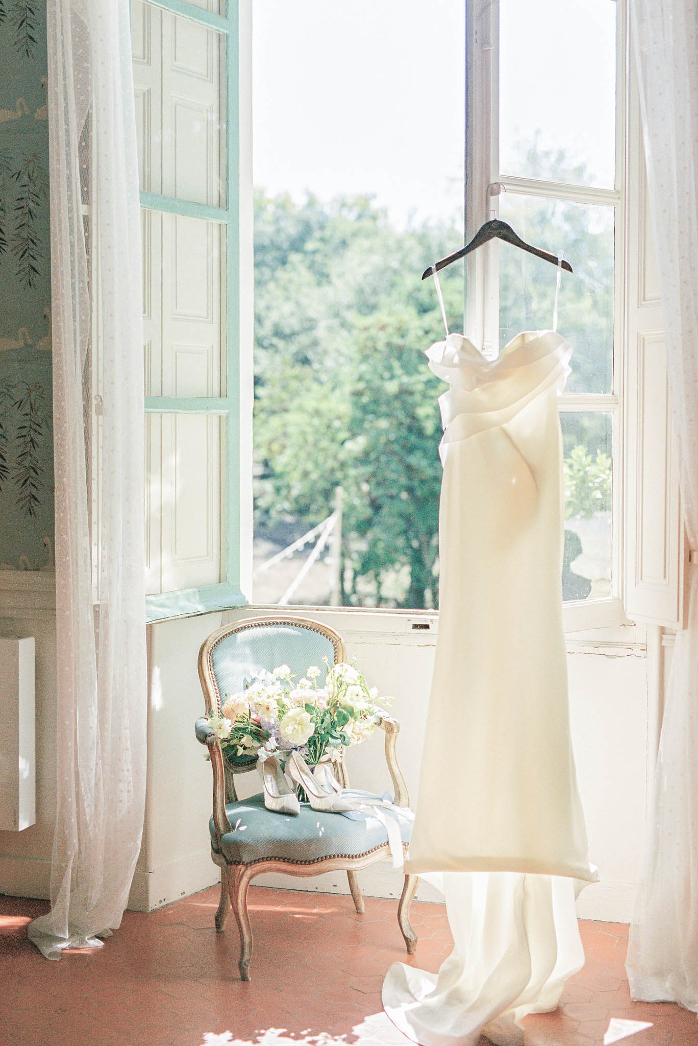 A bridal getting-ready detail shot taken indoors in what appears to be a French château room. An ivory, sleeveless column-style wedding dress with a draped or ruffled neckline detail hangs from a dark wooden hanger on an open white-painted shutter window. A Louis XV-style gilded armchair upholstered in dusty blue-green fabric sits beneath the window, holding a bridal bouquet of white and blush blooms — likely peonies and ranunculus with greenery — alongside a pair of strappy ivory heeled sandals. The room features pale blue-green painted shutters, sheer white curtains, and wallpaper with a delicate botanical print, giving the space a classic French interior aesthetic. The composition is a medium-wide interior shot with natural backlight streaming through the open window.