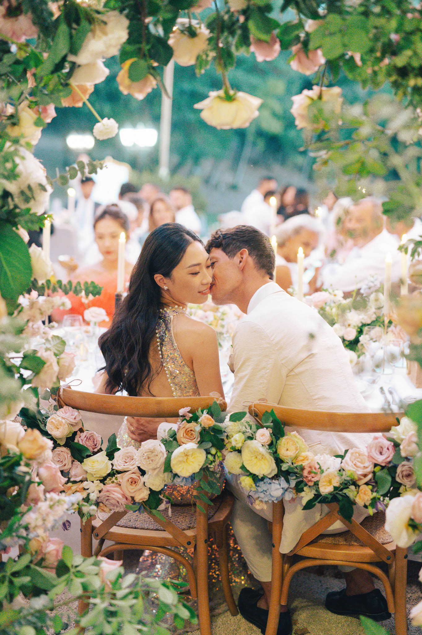 Groom kissing bride at sweetheart table decorated with blush roses and garden roses during outdoor evening reception