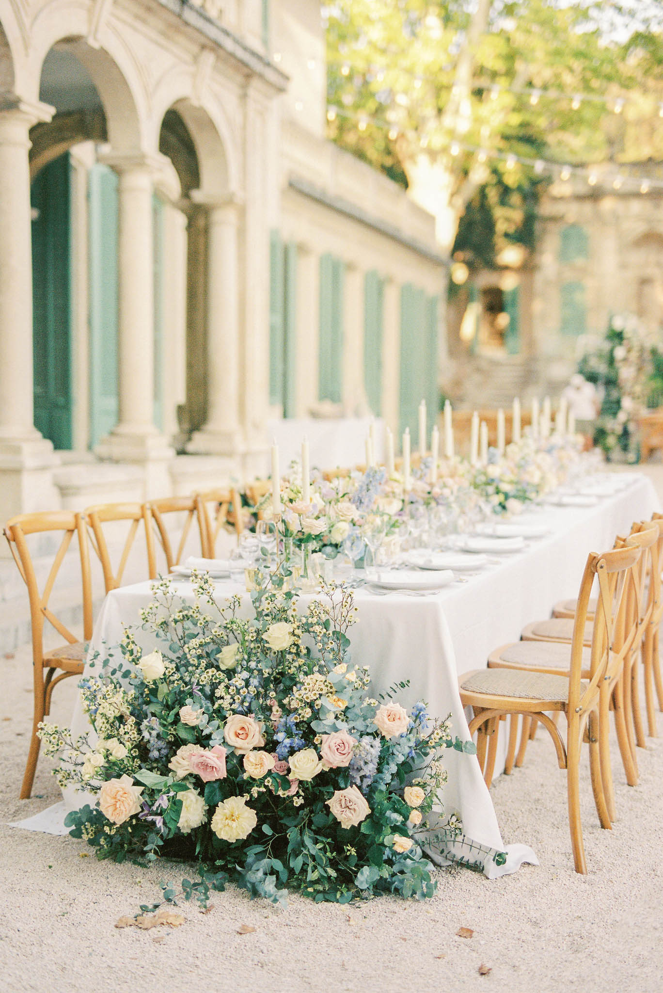 Long banquet table with blush and yellow floral centerpieces, taper candles, and string lights in chateau courtyard
