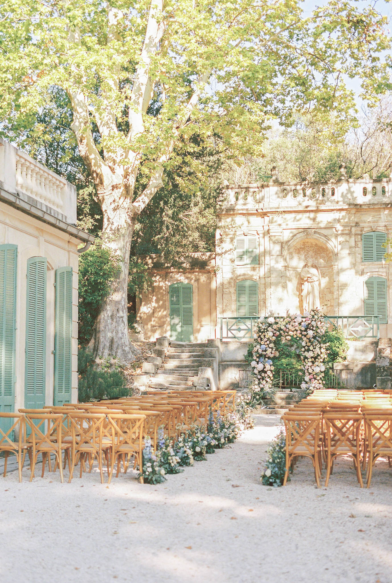 Outdoor ceremony setup with cross-back chairs and floral arch in chateau courtyard with sage green shutters