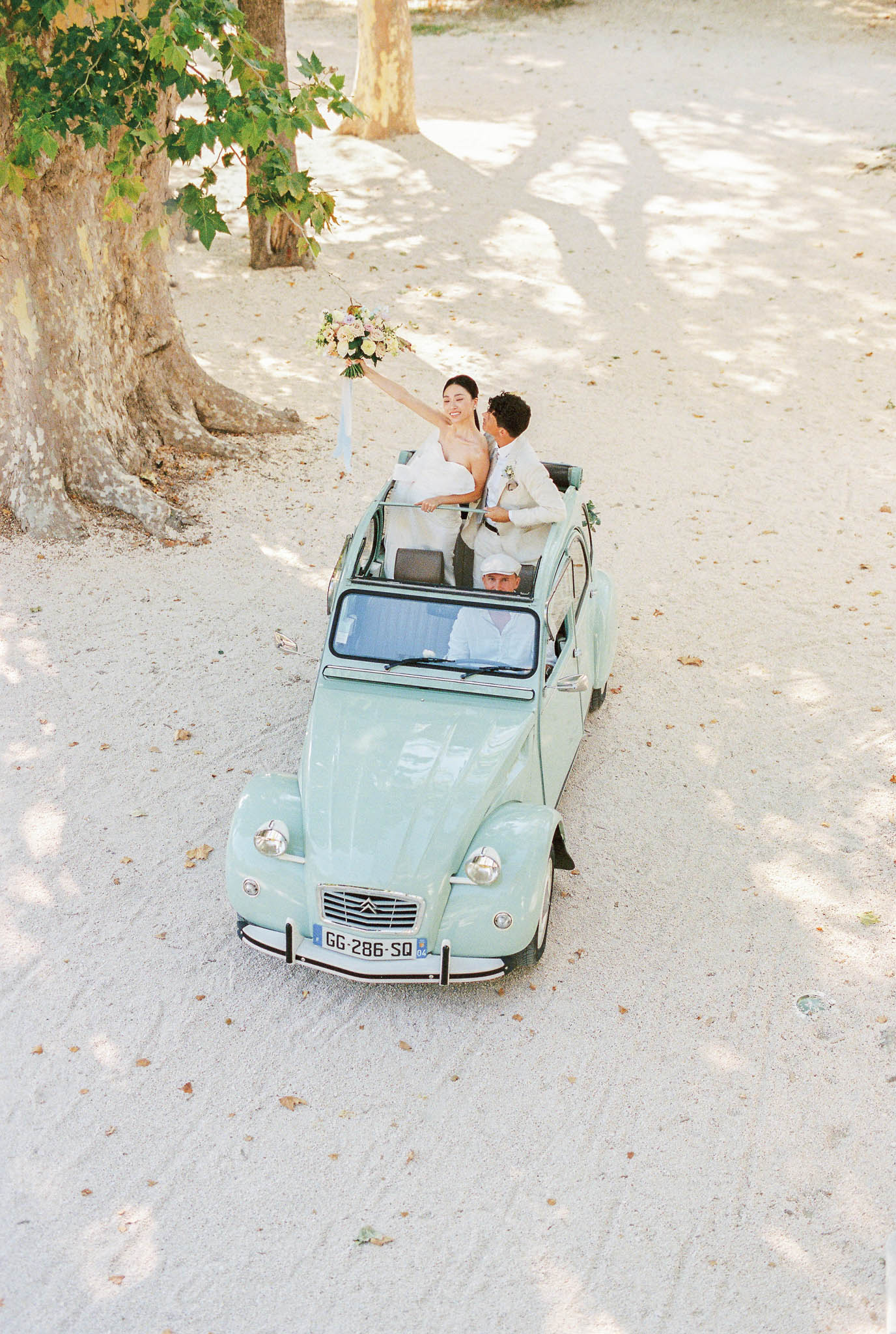The bride and groom are riding in the back of a vintage mint-green Citroën 2CV with the soft top open, driven by a chauffeur wearing a white shirt and cap. The shot is taken from an elevated angle looking down onto a pale gravel driveway lined with large plane trees. The bride, wearing a white strapless gown, raises her bouquet of soft pink, ivory, and blush blooms with trailing white ribbons in a celebratory gesture while smiling broadly; the groom, dressed in a light beige suit with a boutonnière, leans toward her. The composition is a wide overhead portrait capturing the full car and the dappled light across the gravel surface.