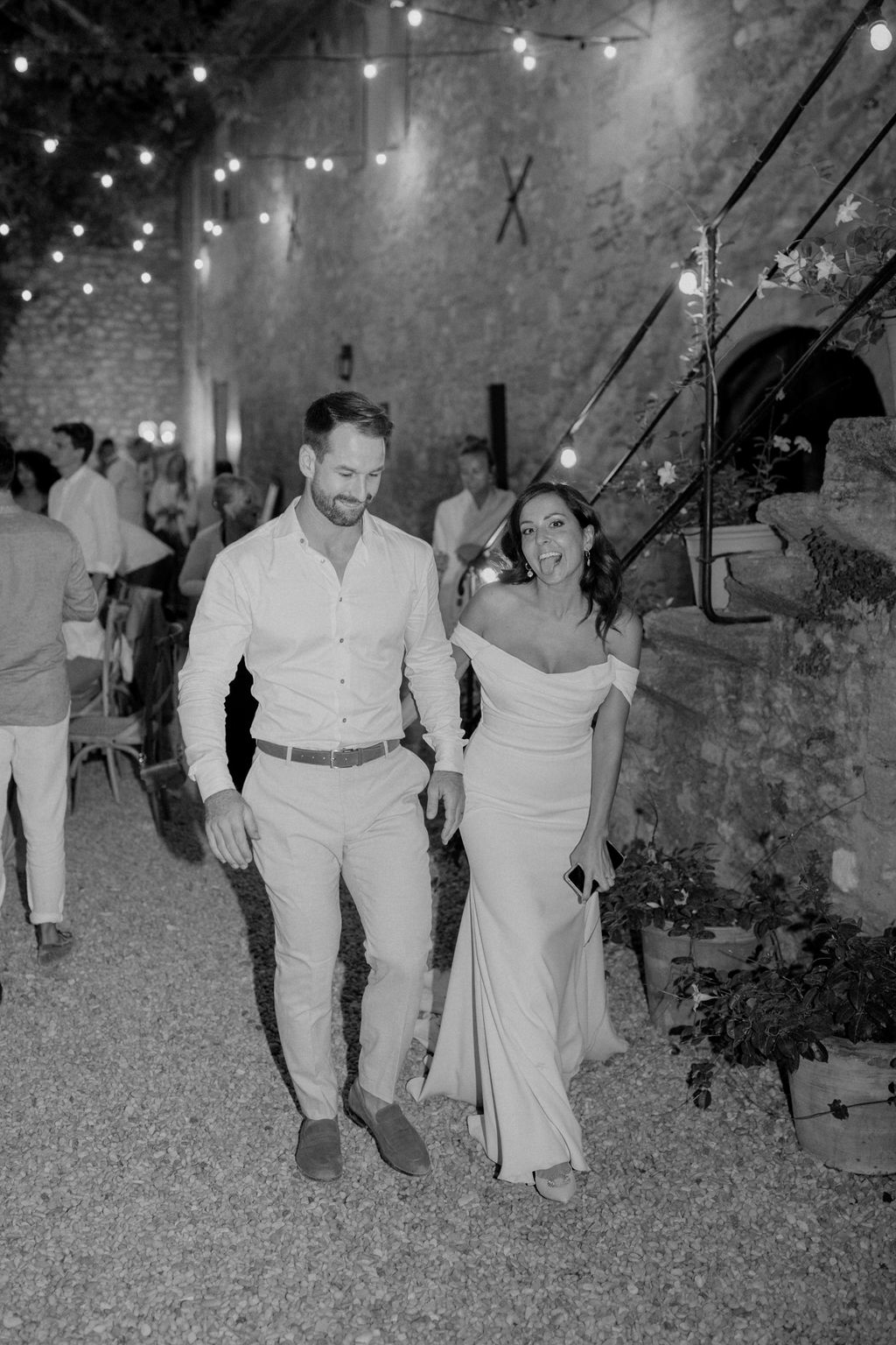This black-and-white image captures a couple walking together during what appears to be an outdoor evening reception at a French stone venue, likely a château or mas. The woman wears a form-fitting off-the-shoulder gown and is laughing playfully toward the camera, while the man beside her wears a casual open-collar white shirt and light trousers, smiling downward. The setting is a gravel courtyard flanked by exposed stone walls, with string globe lights strung overhead creating bright, high-contrast highlights against the dark evening sky. In the background, approximately 15–20 guests dressed predominantly in white and light tones are seated or standing at what appears to be a long dining table, with arched stone doorways and a staircase with climbing florals visible to the right. The image has strong tonal contrast with deep blacks in the shadows and crisp whites in the lighting, giving it a candid, documentary feel. The overall styling is relaxed and warm, consistent with a rustic yet refined outdoor summer reception.