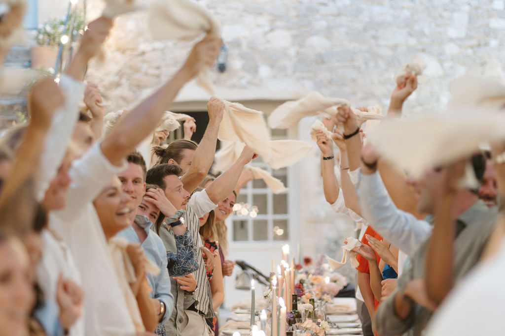 A lively outdoor reception dinner scene where a large group of approximately 20 or more guests seated along a long banquet table are enthusiastically waving cream and ivory linen napkins in the air, a tradition commonly seen at French weddings. The table is styled with tall taper candles in warm tones, low floral arrangements featuring peach, coral, and mixed blooms, and laid place settings. The setting is an outdoor courtyard against a rustic stone building with white-framed windows, suggesting a French country property. Guests are dressed in casual to smart-casual attire in a mix of blues, grays, and prints, and the overall decor palette is warm and natural with cream linens and candlelight. The shot is taken from a low, mid-range angle along the length of the table, giving a dynamic perspective of the raised arms and waving napkins.