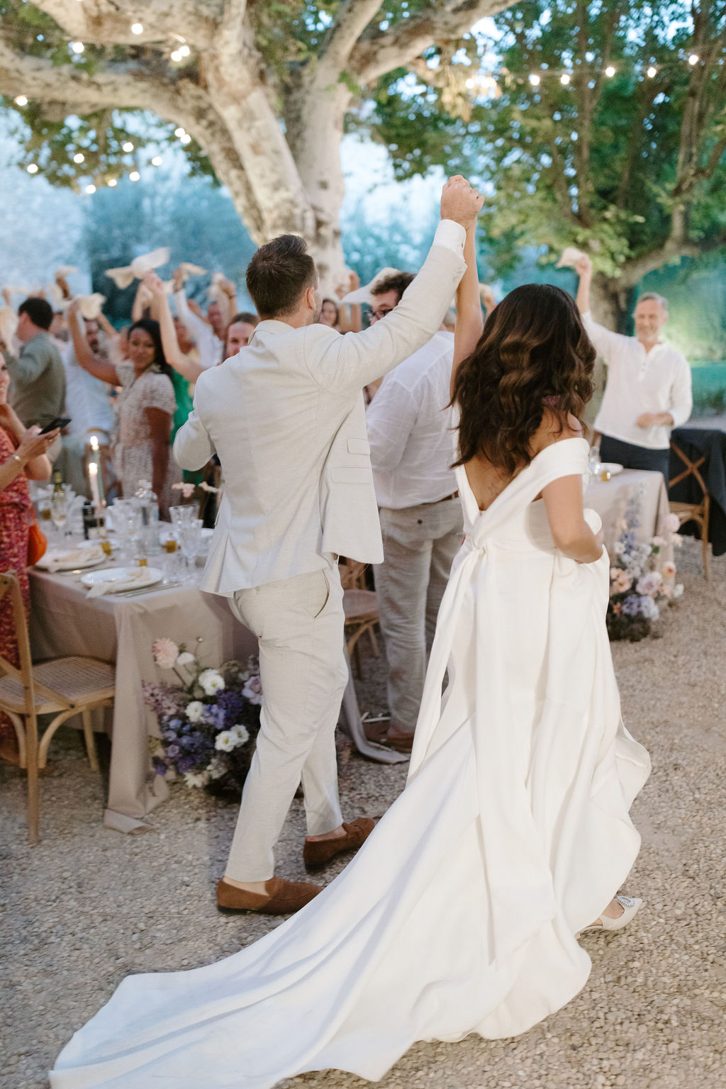 Bride and groom make celebratory entrance to outdoor reception under fairy lights as guests wave napkins