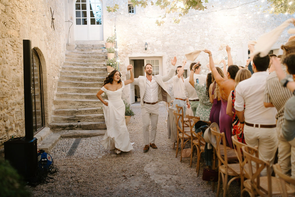 Couple making celebratory entrance arms raised guests cheering in rustic stone mas courtyard with string lights