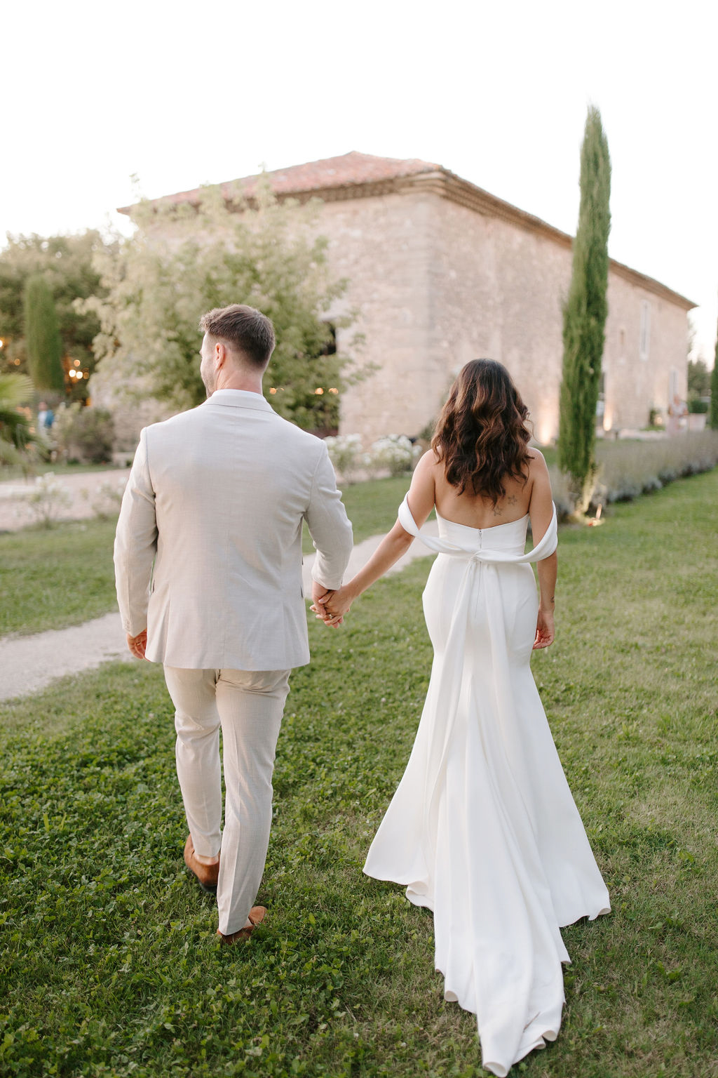 Bride and groom walking hand-in-hand toward Provencal stone venue at golden hour from behind