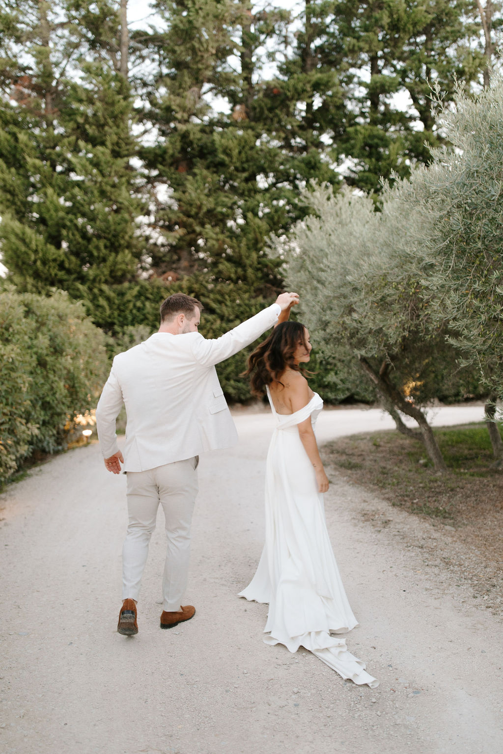 Groom spinning bride on olive tree-lined gravel path, both in white, viewed from behind