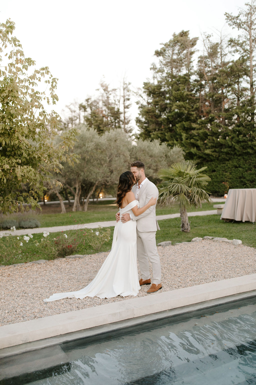 Bride in off-shoulder gown and groom in sand suit kiss beside rectangular pool in landscaped garden