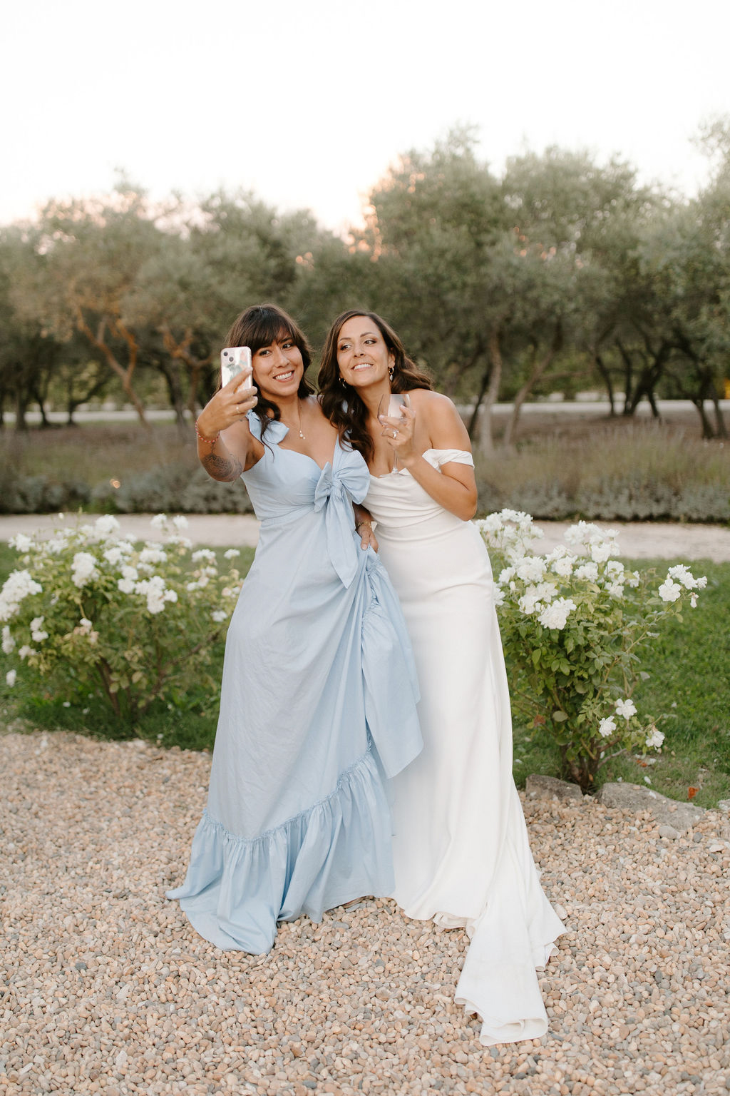 Bride in ivory gown and companion in powder-blue dress take selfie on gravel path with olive trees