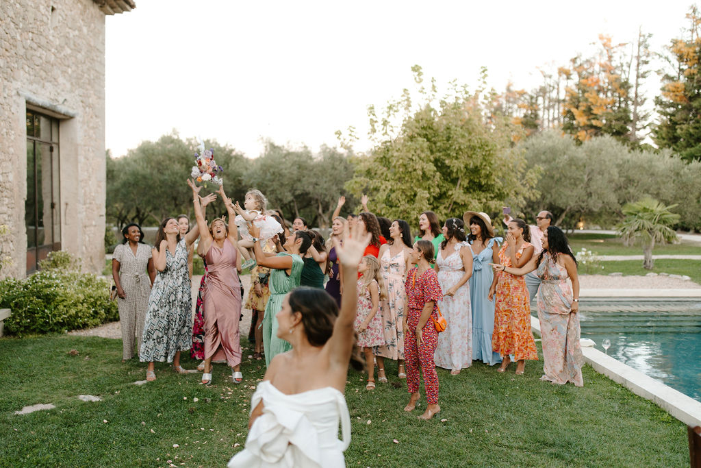 The bride, visible from behind in a white off-the-shoulder dress, is tossing her bouquet — a compact arrangement with colorful blooms including what appears to be coral, pink, and purple flowers — toward a group of approximately 20 female guests gathered on a lawn. The guests are reaching upward with arms raised, expressions animated with excitement. The group is dressed in a wide variety of colorful outfits including dusty rose, sage green, red, light blue, orange floral, and patterned wrap dresses, creating a vibrant and eclectic palette. The setting is an outdoor garden area beside a rectangular swimming pool, adjacent to a stone building with large steel-framed windows, consistent with a Provençal mas or bastide-style venue. The shot is taken from behind the bride at medium distance, capturing both her and the full group in a wide portrait-style frame during what appears to be late afternoon golden hour light.