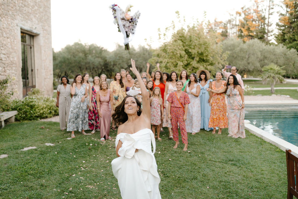 Bride in off-the-shoulder gown tossing her bouquet to guests on the lawn of a French stone estate