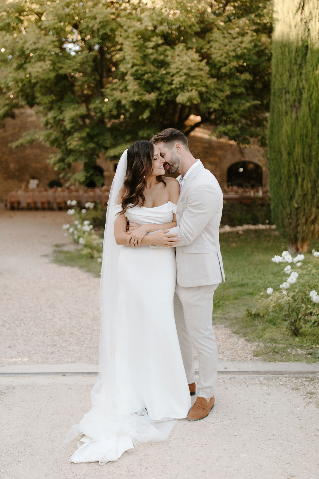 Bride in off-shoulder satin gown and groom in sand-beige suit embracing on a gravel path at a French domaine
