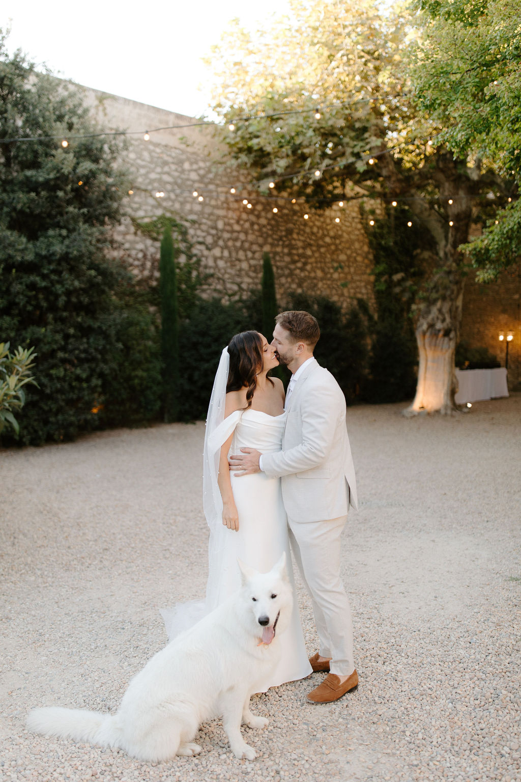 Couple kissing at golden hour with white Swiss Shepherd dog at feet under courtyard string lights