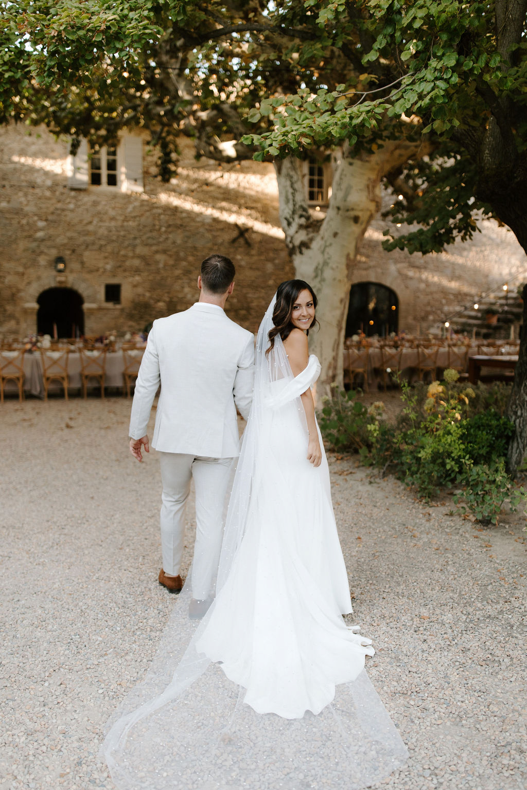 Couple walking from camera at bastide courtyard bride turning back with cathedral veil reception tables behind