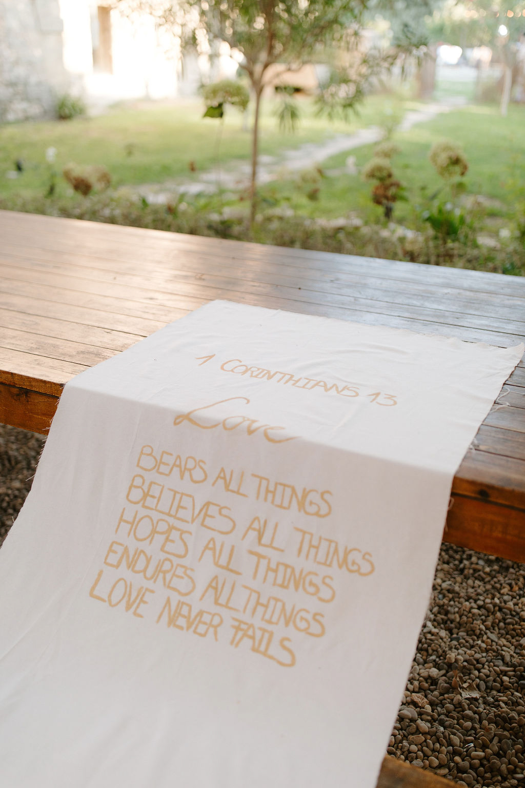 A close-up detail shot of a white fabric table runner laid across a rustic wooden farm table in an outdoor setting. The runner features hand-lettered text in golden yellow ink, displaying the biblical verse 1 Corinthians 13: 'Love bears all things, believes all things, hopes all things, endures all things, love never fails.' The lettering combines a mix of cursive script for the word 'Love' and block capitals for the remaining lines. The table is set on a gravel surface with a garden visible in the soft-focus background. The styling reflects a rustic, faith-inspired wedding aesthetic with a warm neutral color palette of ivory and gold.