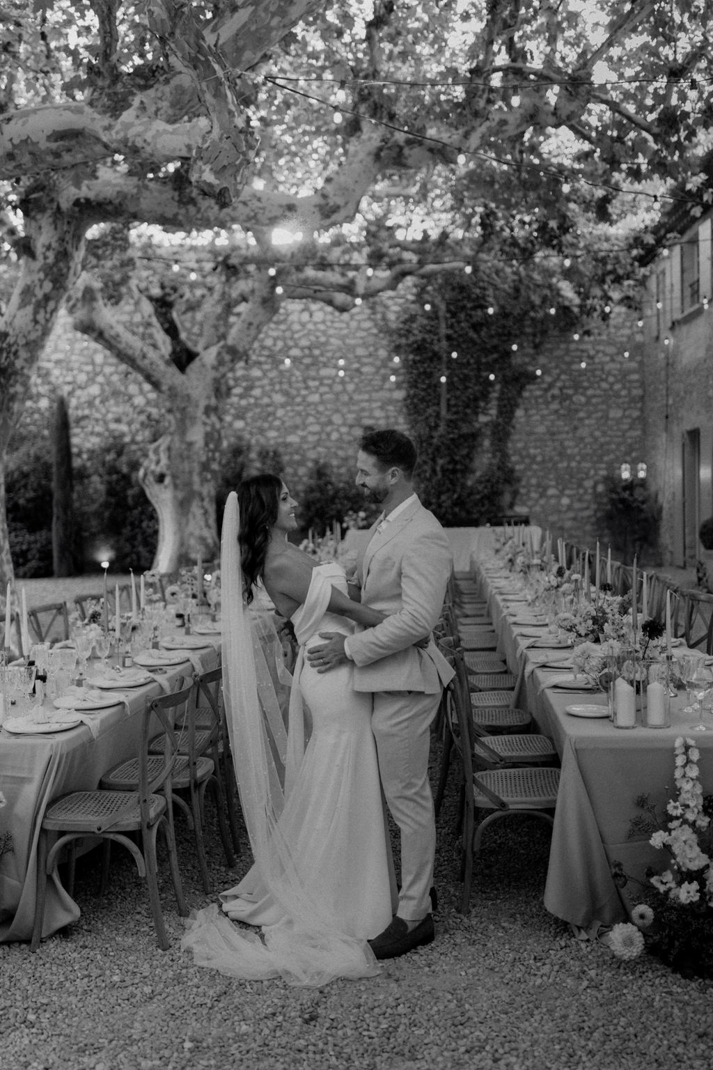 Black and white portrait of bride and groom embracing between banquet tables under plane trees with string lights