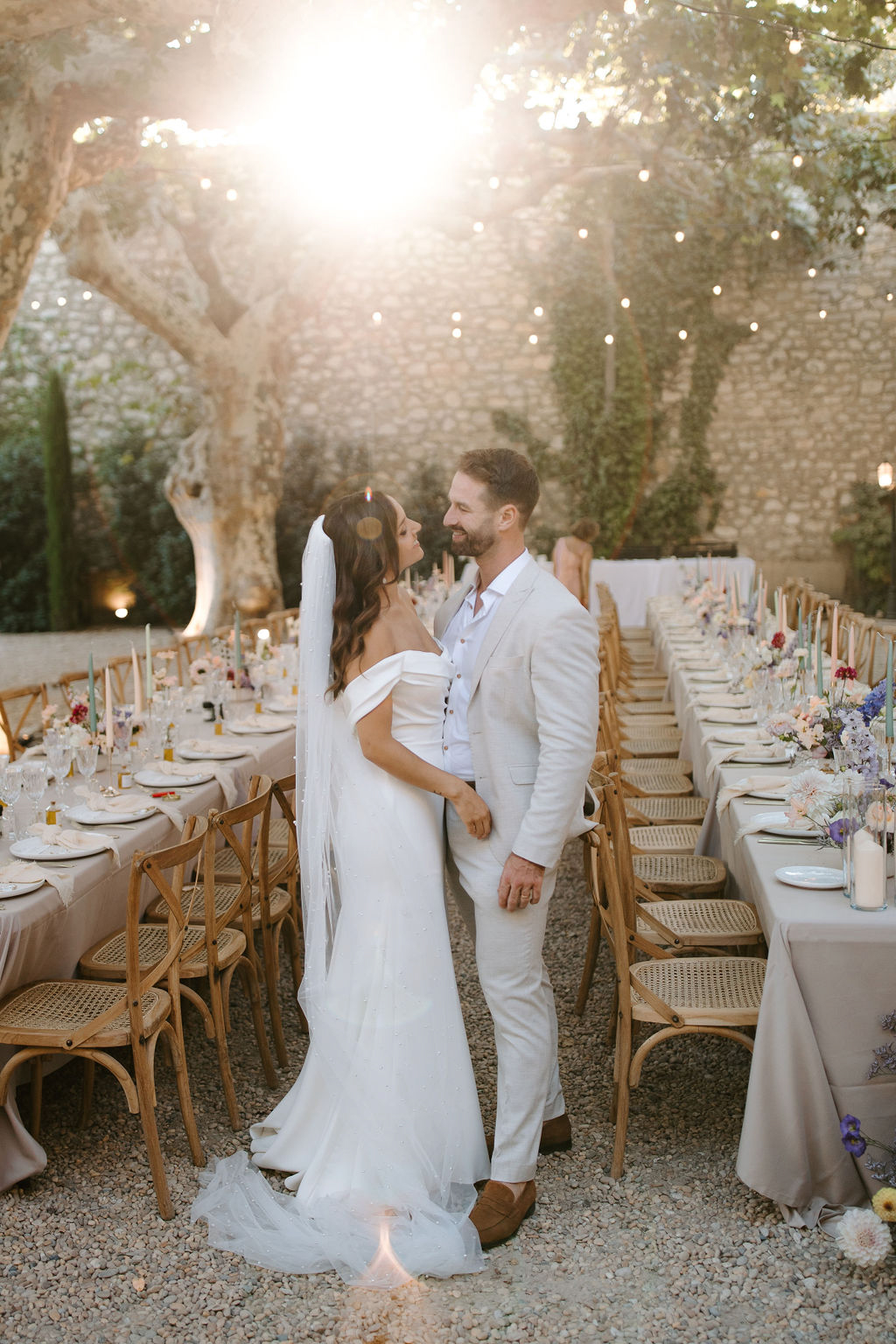 Bride in off-shoulder gown and groom in cream suit between outdoor reception tables with golden backlight