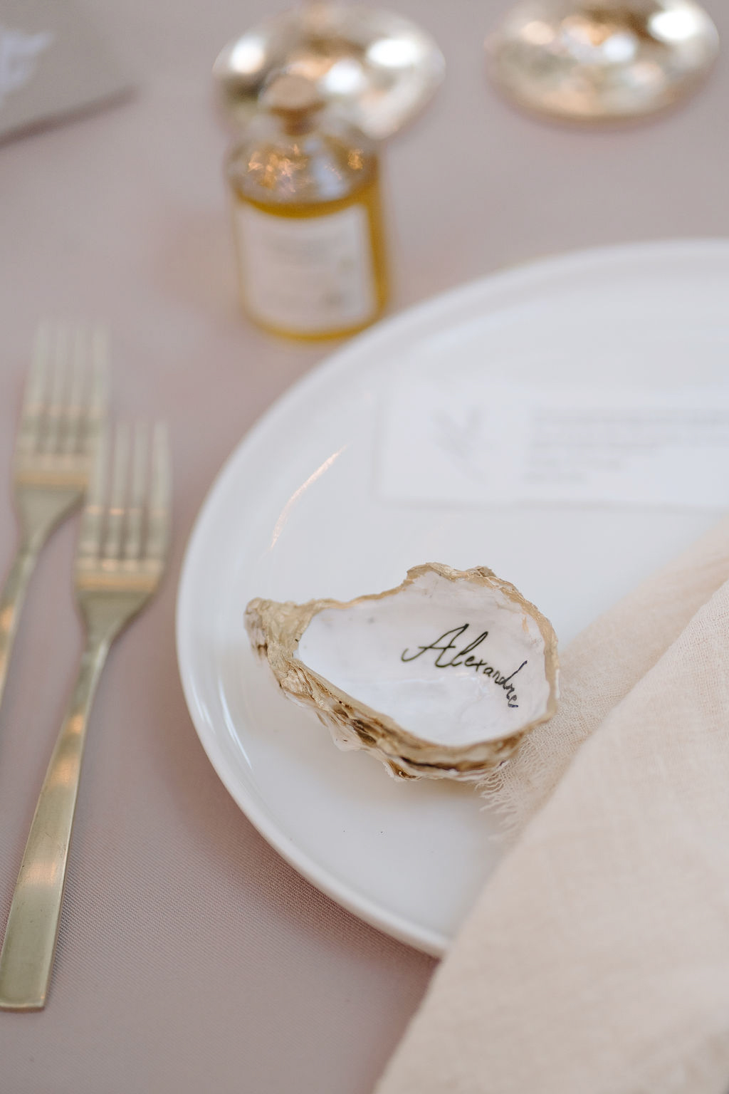 Oyster shell escort card with black calligraphy on white plate with blush linen gold flatware and honey favour