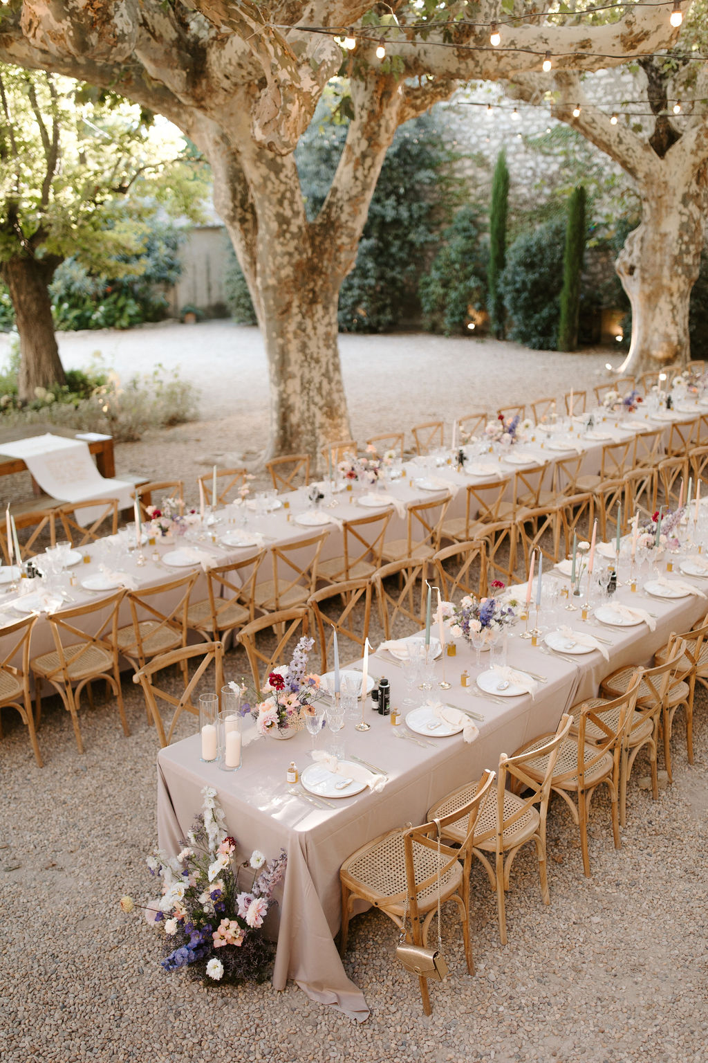 Two long reception tables with mauve linens and cross-back chairs under plane trees with wildflower centerpieces and strin...