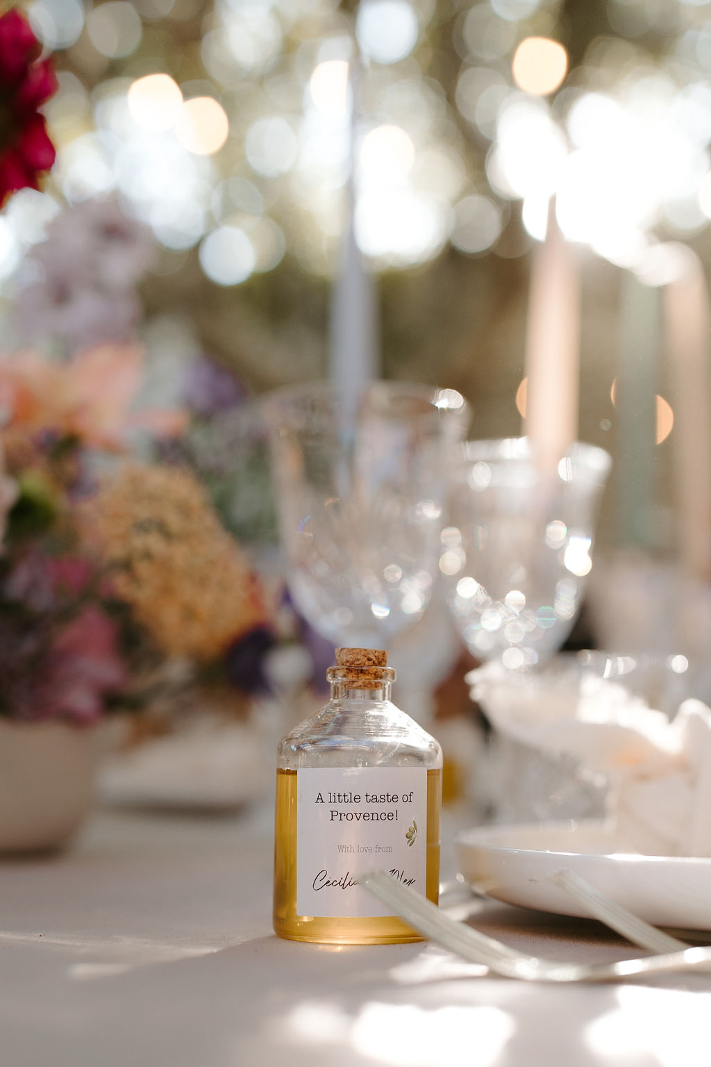 Close-up detail shot of a personalized wedding favor placed on a reception table. The favor is a small cork-stoppered glass bottle filled with golden-yellow liquid — likely olive oil or honey — labeled 'A little taste of Provence! With love from Cecilia & Max.' In the blurred background, crystal wine glasses, tall taper candles in blush and grey tones, white plates, and a lush floral centerpiece featuring pink, crimson, peach, and lavender blooms are visible. The table is covered with a white linen, and the overall decor palette combines warm jewel tones with soft neutrals in what appears to be an outdoor Provence-style reception setting.
