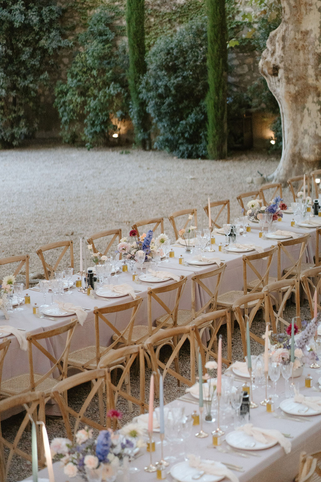 An outdoor wedding reception dinner setup photographed at dusk in a gravel courtyard, likely at a French château or historic estate. Two long banquet tables are dressed in dusty mauve linen tablecloths and lined with natural wood cross-back chairs with rattan seats. Each place setting includes white plates, folded linen napkins, multiple crystal glasses, and small amber glass favor bottles. Floral centerpieces feature loose arrangements of blue delphiniums, blush ranunculus, white daisies, and deep pink dahlias placed at intervals along the tables, accompanied by slender taper candles in pastel tones of blush, sage, and ivory. The image is a wide angled shot taken from a slightly elevated perspective looking down the length of the tables, with lit ground-level spotlights illuminating cypress trees and ivy-covered walls in the background. Potential venue feature image.