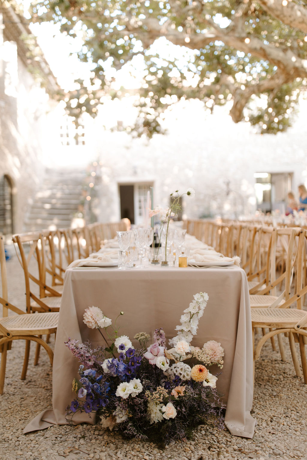 An outdoor reception dinner setup in a courtyard with a gravel floor, surrounded by stone walls and a large tree with fairy lights strung through its canopy overhead. The foreground features the end of a long rectangular feasting table draped in a taupe linen tablecloth, with a loose cascading floral arrangement placed at the base of the table on the ground, composed of blue delphiniums, blush carnations, white lisianthus, lavender blooms, ivory stock flowers, and small filler flowers in a wildflower style. The table is set with crystal glassware, white folded napkins, and small bud vases with delicate single-stem flowers including cosmos and pale pink blooms, along with gold votive candle holders. Natural wood cross-back chairs with rattan seats line both sides of the table. A second table is partially visible in the background with guests seated. The overall decor palette is soft and muted — taupe, blush, lilac, and dusty blue — with a relaxed, garden-party aesthetic. Wide shot taken from ground level at the table end. Potential venue feature image.