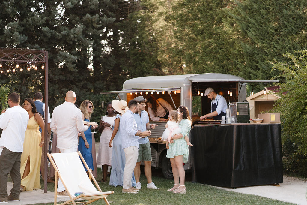 Guests gathered around vintage silver food truck with festoon lights at garden cocktail hour